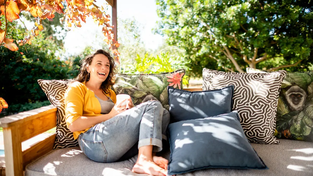Young woman laughing while relaxing outside on her patio sofa and drinking a cup of coffe