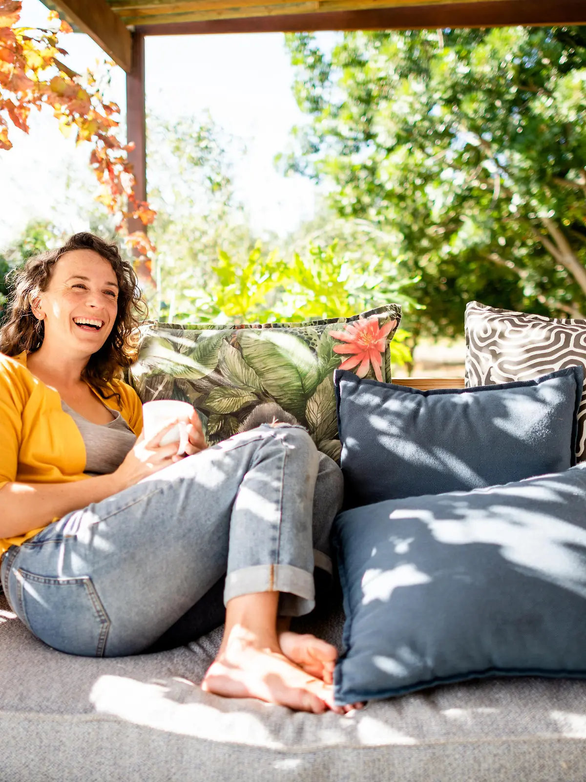 Young woman laughing while relaxing outside on her patio sofa and drinking a cup of coffe