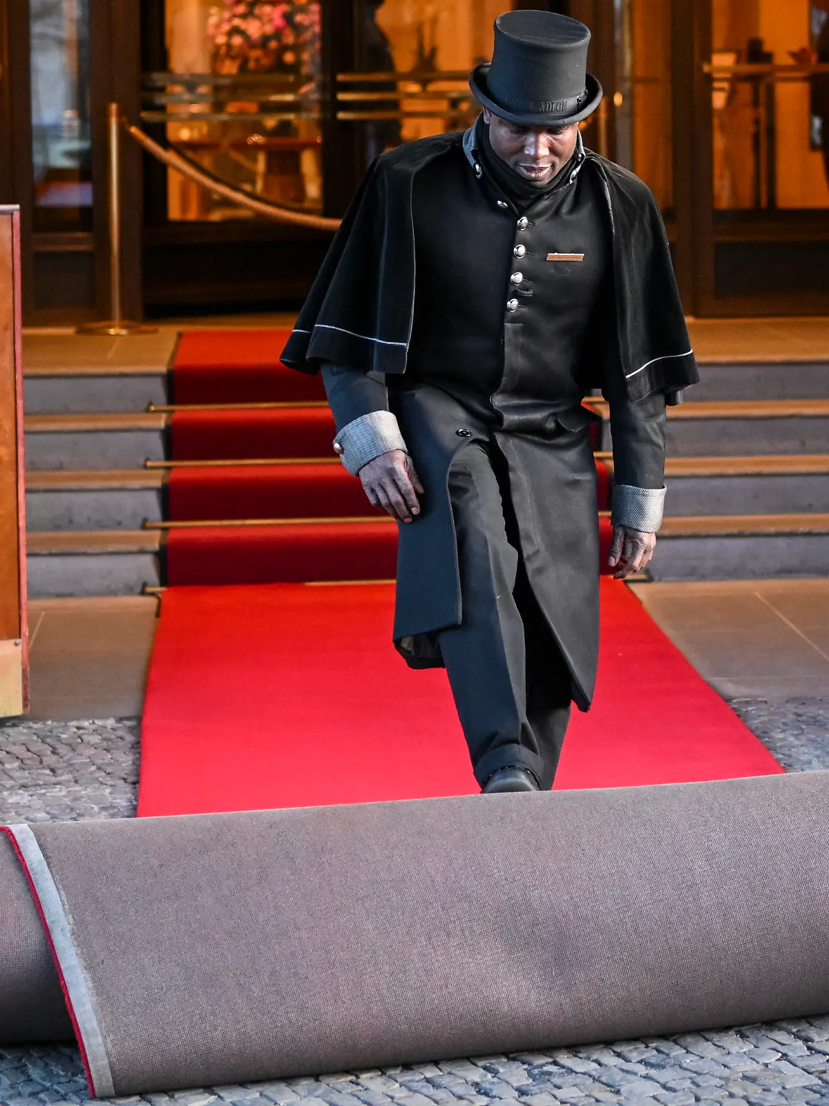 PRODUKTION - 16.03.2023, Berlin: Doorman Timothy Uhunamure rollt vor dem Hotel Adlon den roten Teppich aus. Zu manchen Details eines königlichen Besuchs wird traditionell geschwiegen. Insidern zufolge wird König Charles III. wohl dort unterkommen, wo bereits seine Mutter Queen Elizabeth II. zu Gast war. Eine der drei Präsidenten-Suiten wurde im Hotel Adlon ihr zu Ehren zur Royal Suite ernannt. Foto: Jens Kalaene/dpa +++ dpa-Bildfunk +++