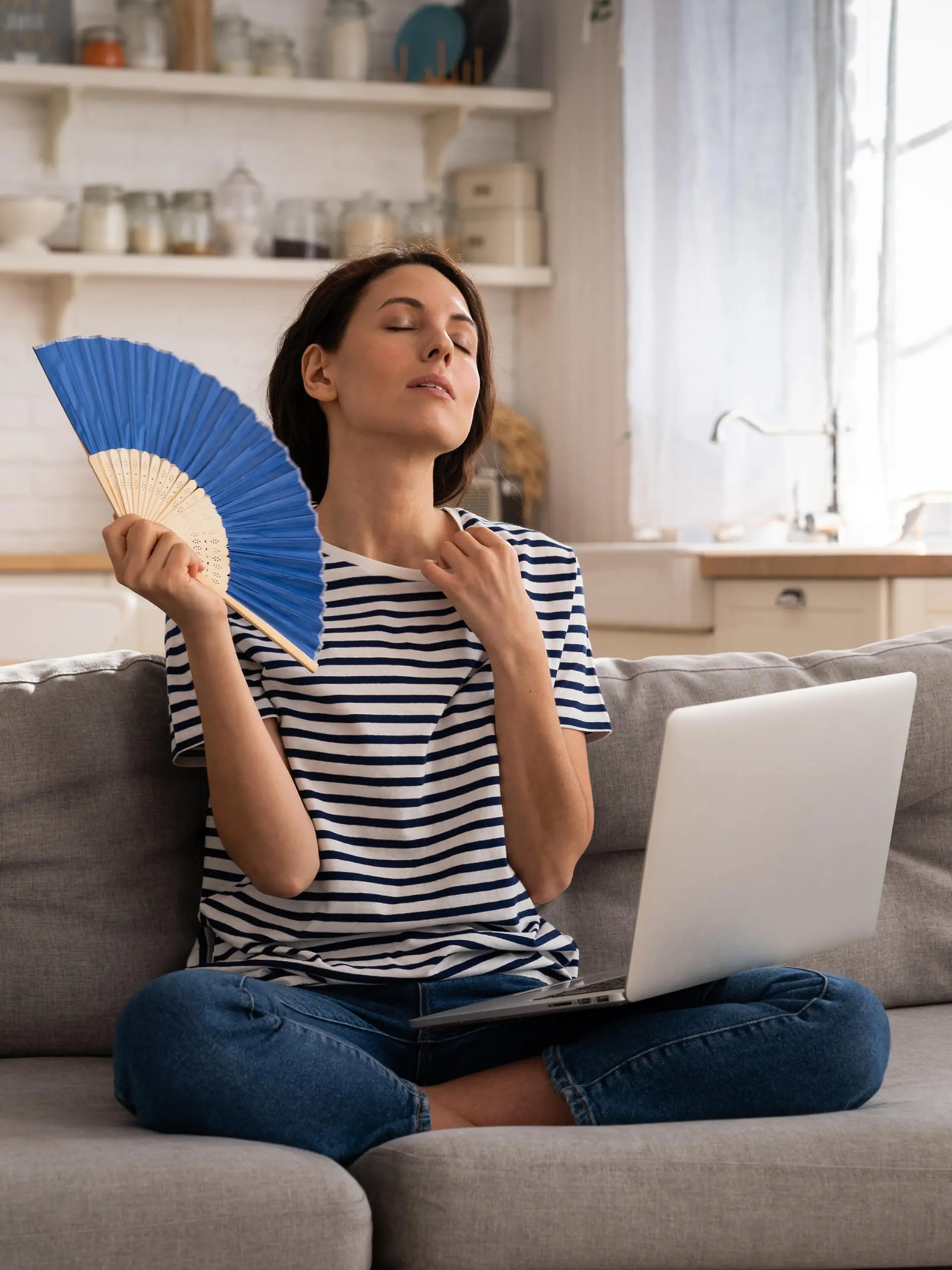 Tired millennial woman suffers from stuffiness and an inoperative air conditioner, waving blue fan sitting on couch at home working on laptop computer. Overheating high temperature, hot summer weather