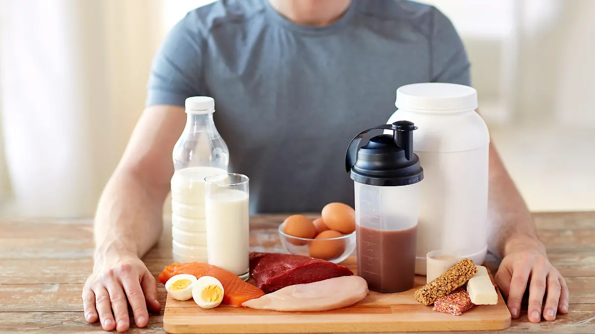 Ein Mann, den man nur halb im Bild zu Gesicht bekommt, sitzt an einem Esstisch. Vor ihm liegt ein Brett mit unterschiedlichen eiweißhaltigen Lebensmitteln, wie Fisch, Proteinshake und Eier.