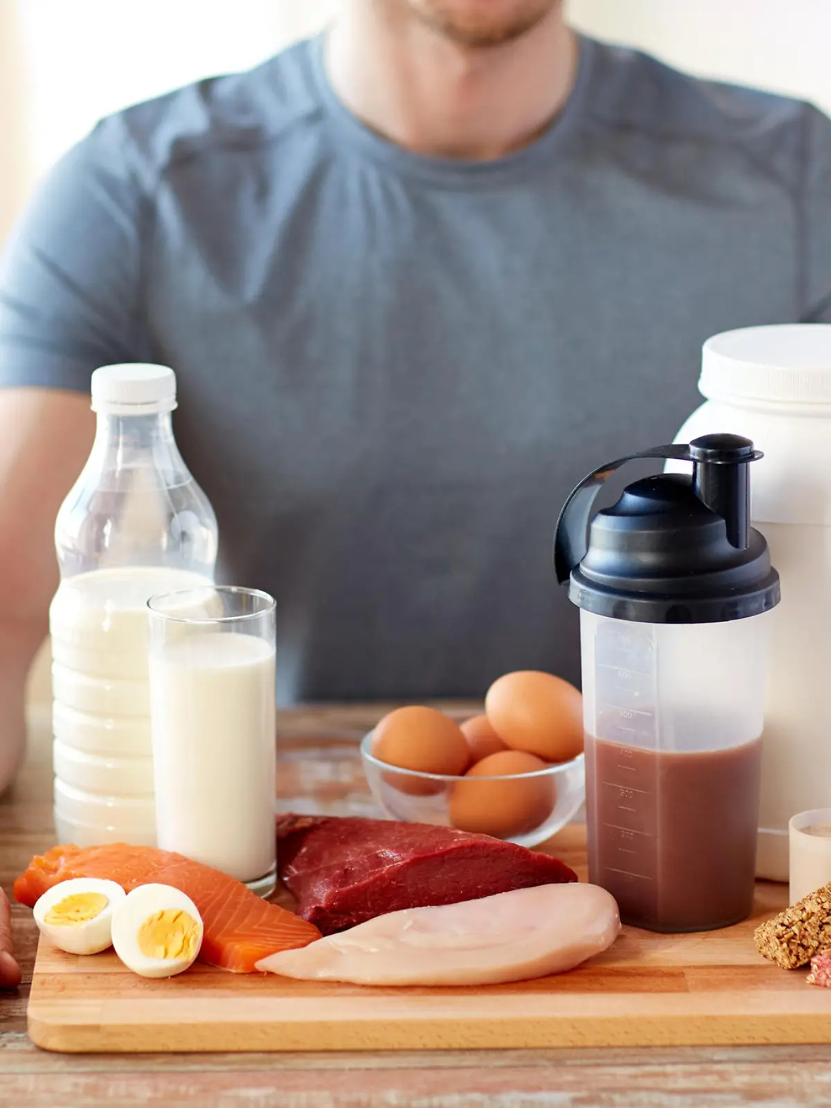 Ein Mann, den man nur halb im Bild zu Gesicht bekommt, sitzt an einem Esstisch. Vor ihm liegt ein Brett mit unterschiedlichen eiweißhaltigen Lebensmitteln, wie Fisch, Proteinshake und Eier.