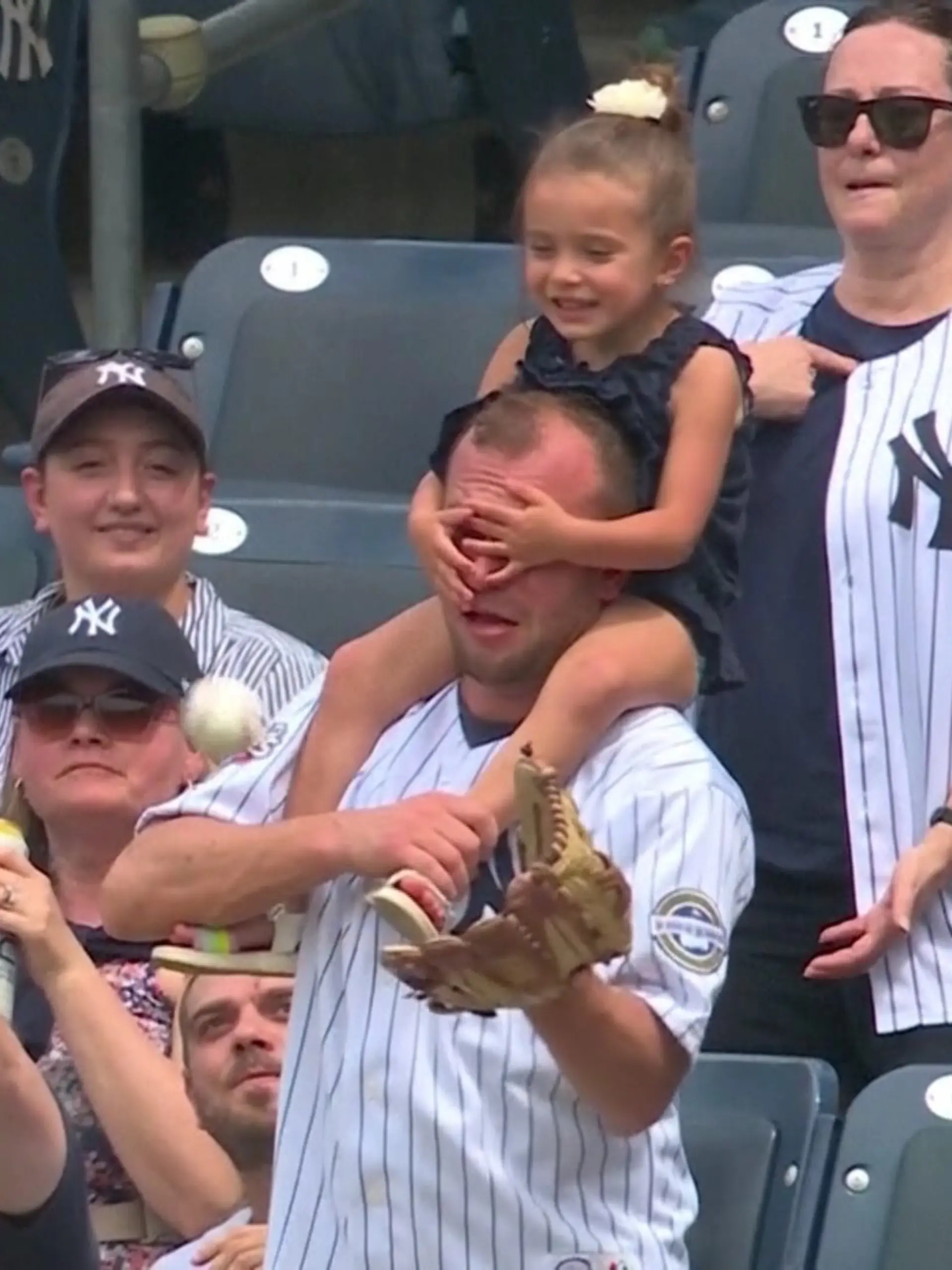 Augen zu, Papa! Ein Mädchen, das auf den Schultern seines Vaters sitz, hält ihm die Augen zu, während der einen Baseball fangen will.