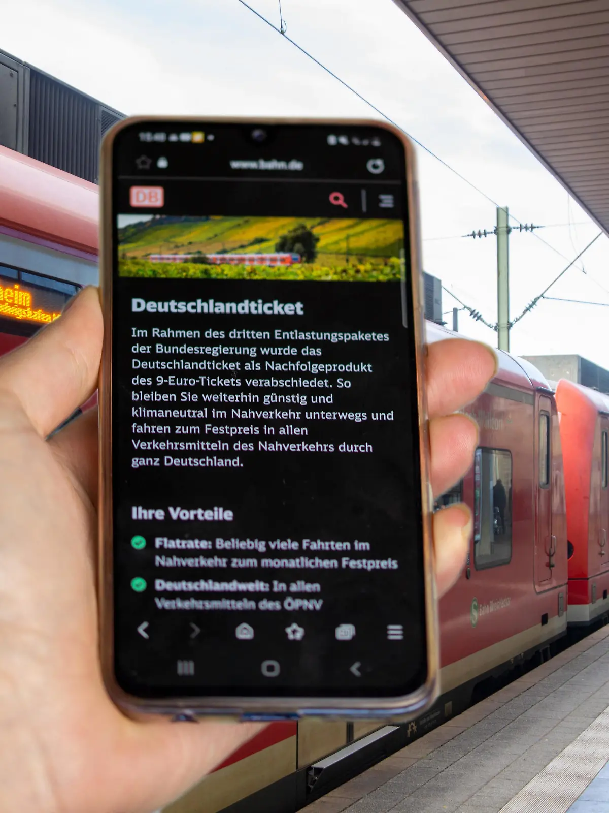 Symbolbild Deutschlandticket, 49-Euro-Ticket: Nahaufnahme eines Smartphones vor ein- und aussteigenden Fahrgästen beim Halt eines Zuges