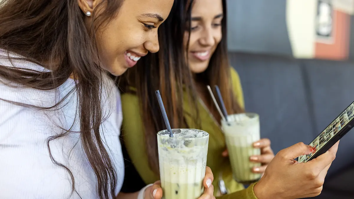 Glückliche Freundinnen sitzen in einem Café und schauen auf ihr Handy, während sie eisgekühlten Matcha Latte trinken.