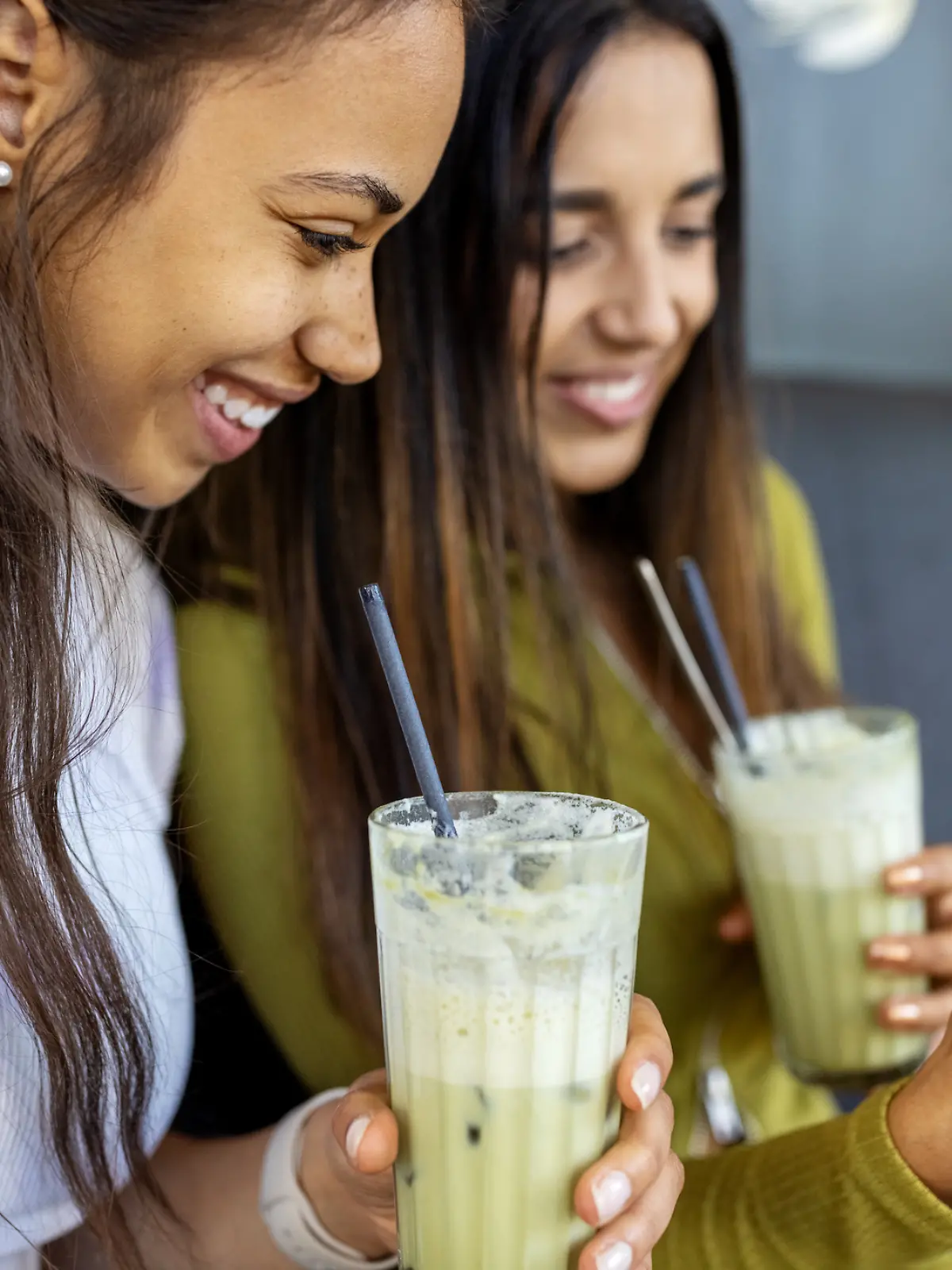 Glückliche Freundinnen sitzen in einem Café und schauen auf ihr Handy, während sie eisgekühlten Matcha Latte trinken.