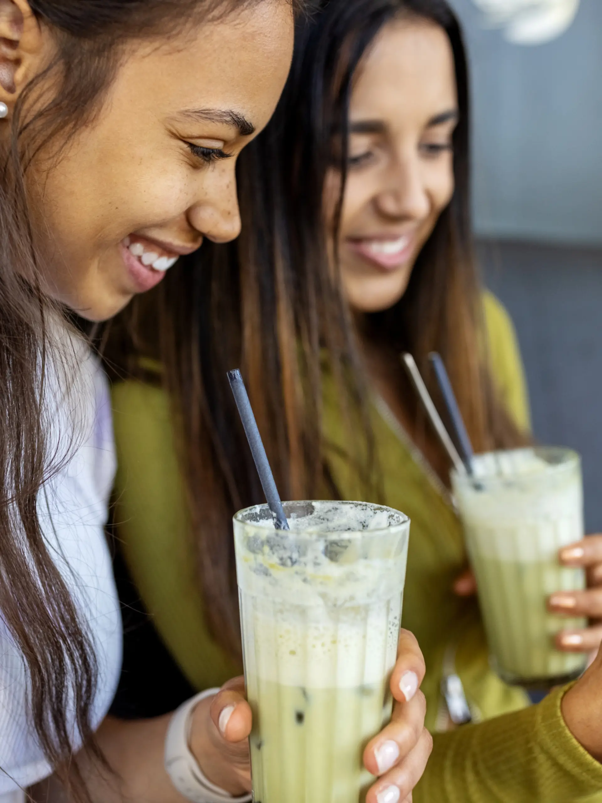 Glückliche Freundinnen sitzen in einem Café und schauen auf ihr Handy, während sie eisgekühlten Matcha Latte trinken.