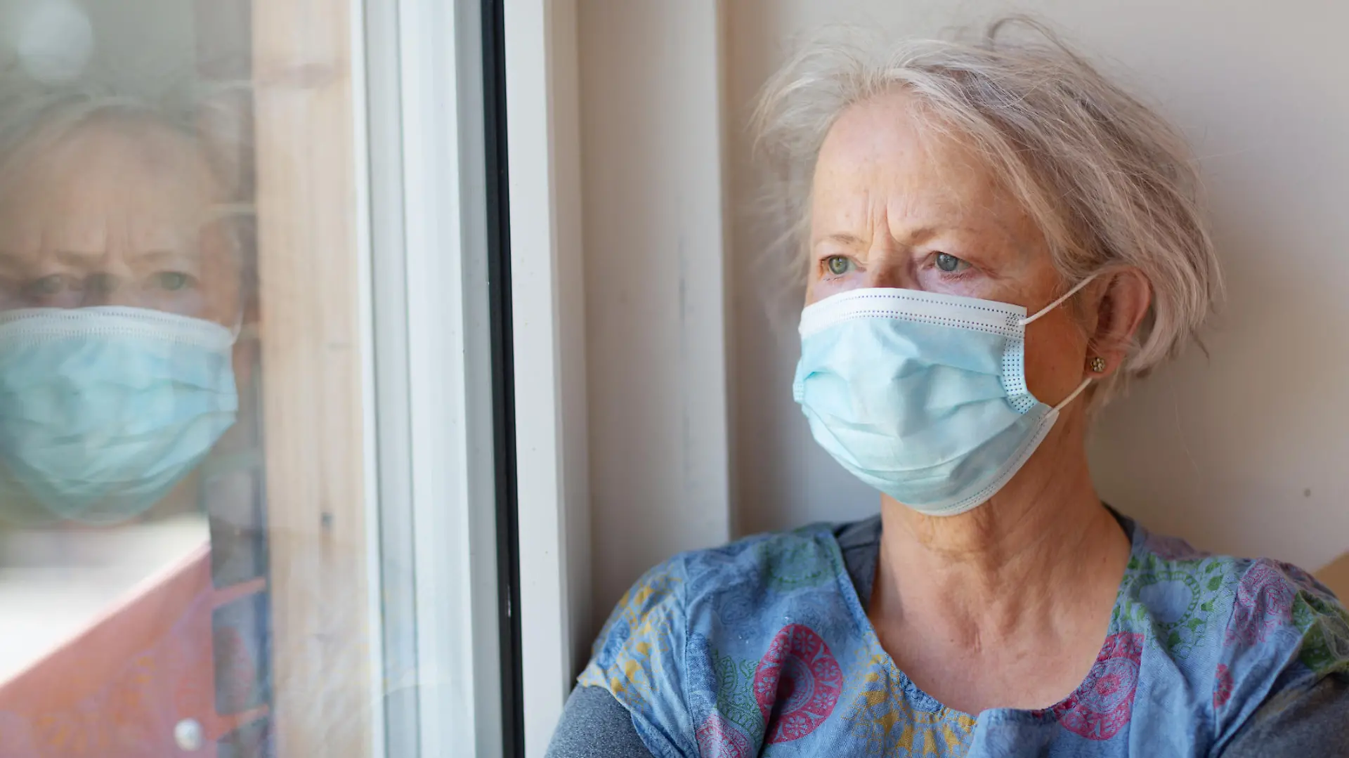 Seniorin mit Maske guckt aus dem Fenster.