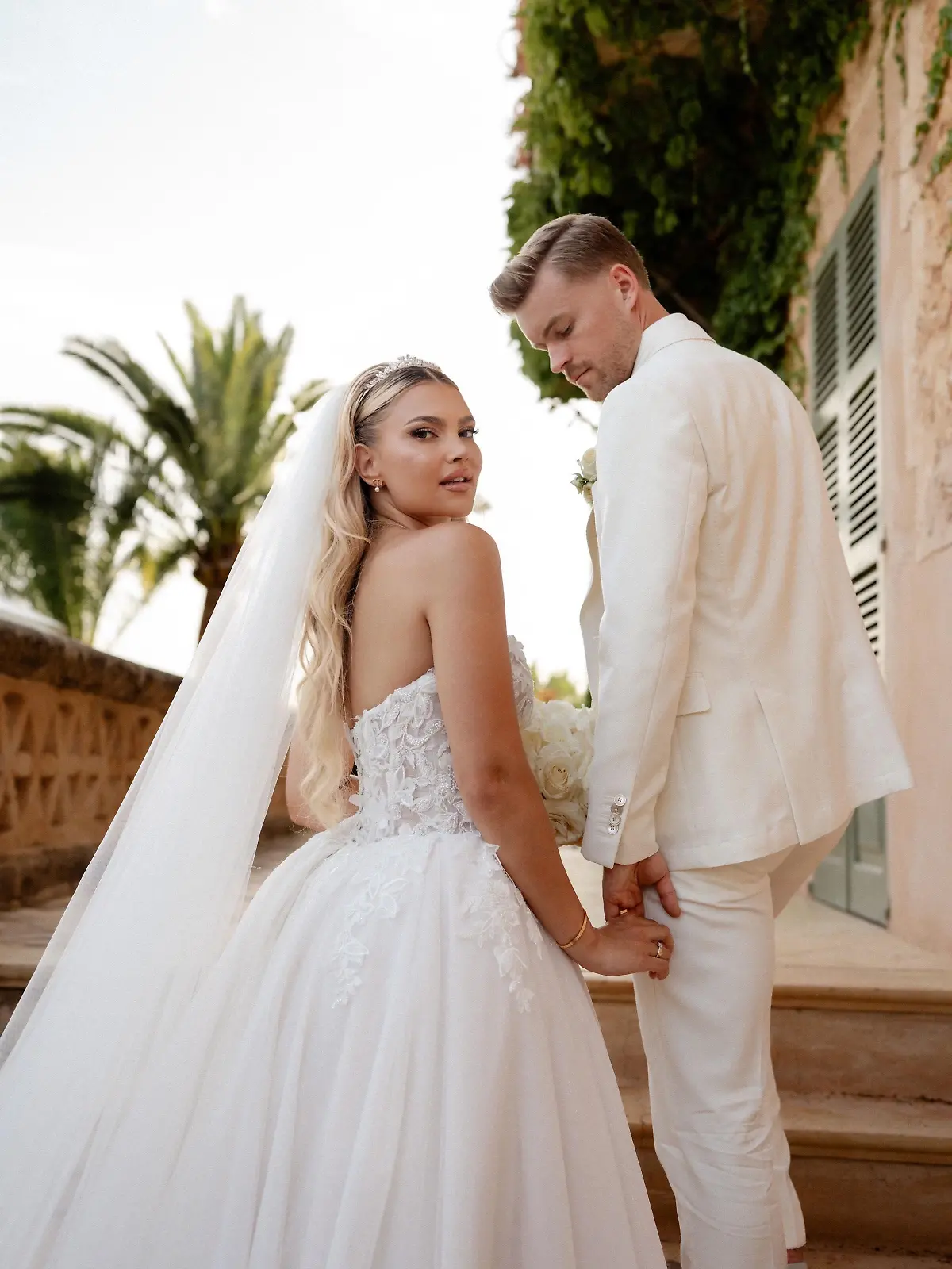 Hochzeitsfoto von Lea und Maximilian Mittelstädt: Beide stehen auf einer Treppe und schauen über ihre Schultern nach hinten - sie in einem weißen Brautkleid, der in einem weißen Anzug