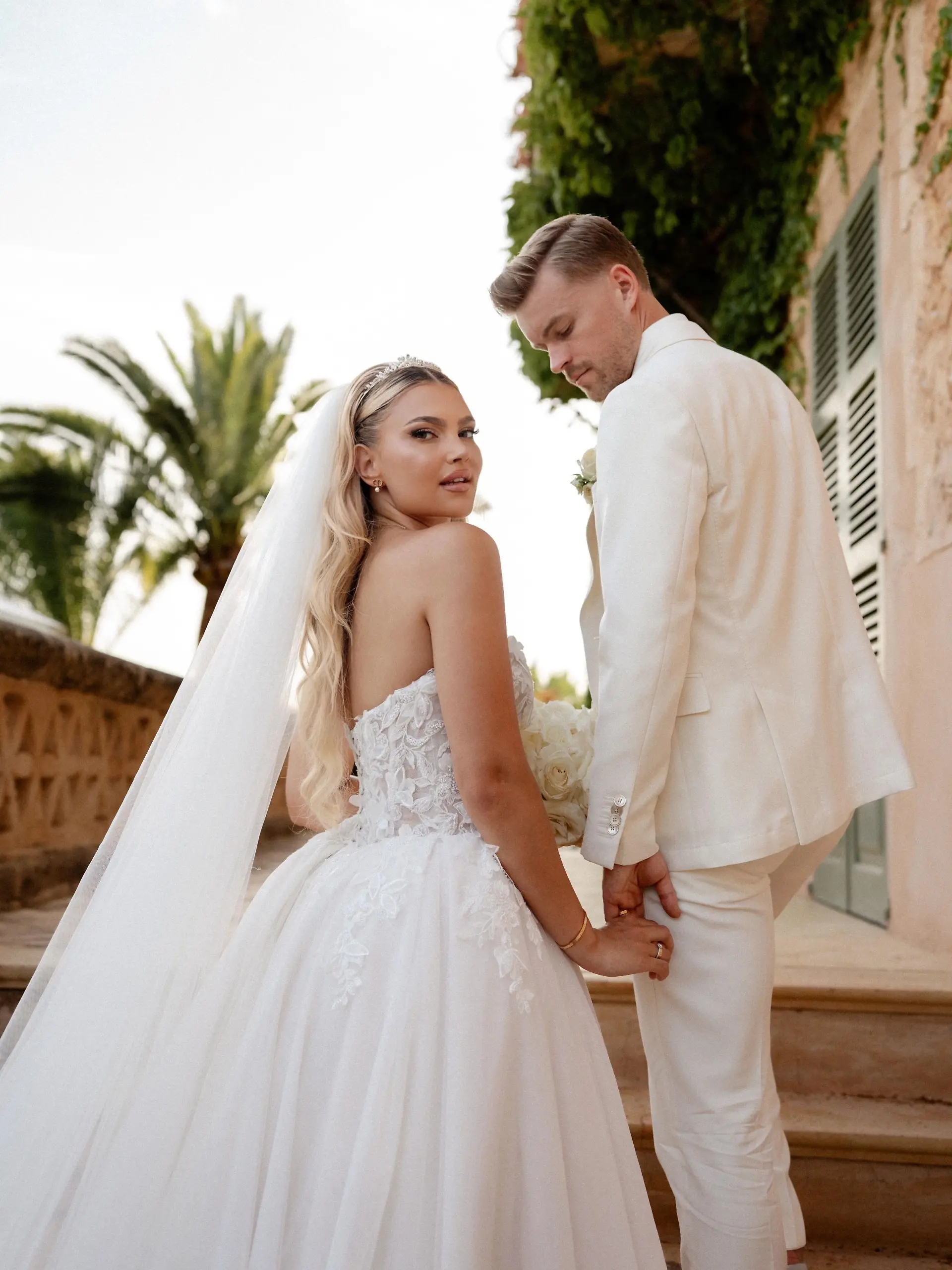 Hochzeitsfoto von Lea und Maximilian Mittelstädt: Beide stehen auf einer Treppe und schauen über ihre Schultern nach hinten - sie in einem weißen Brautkleid, der in einem weißen Anzug