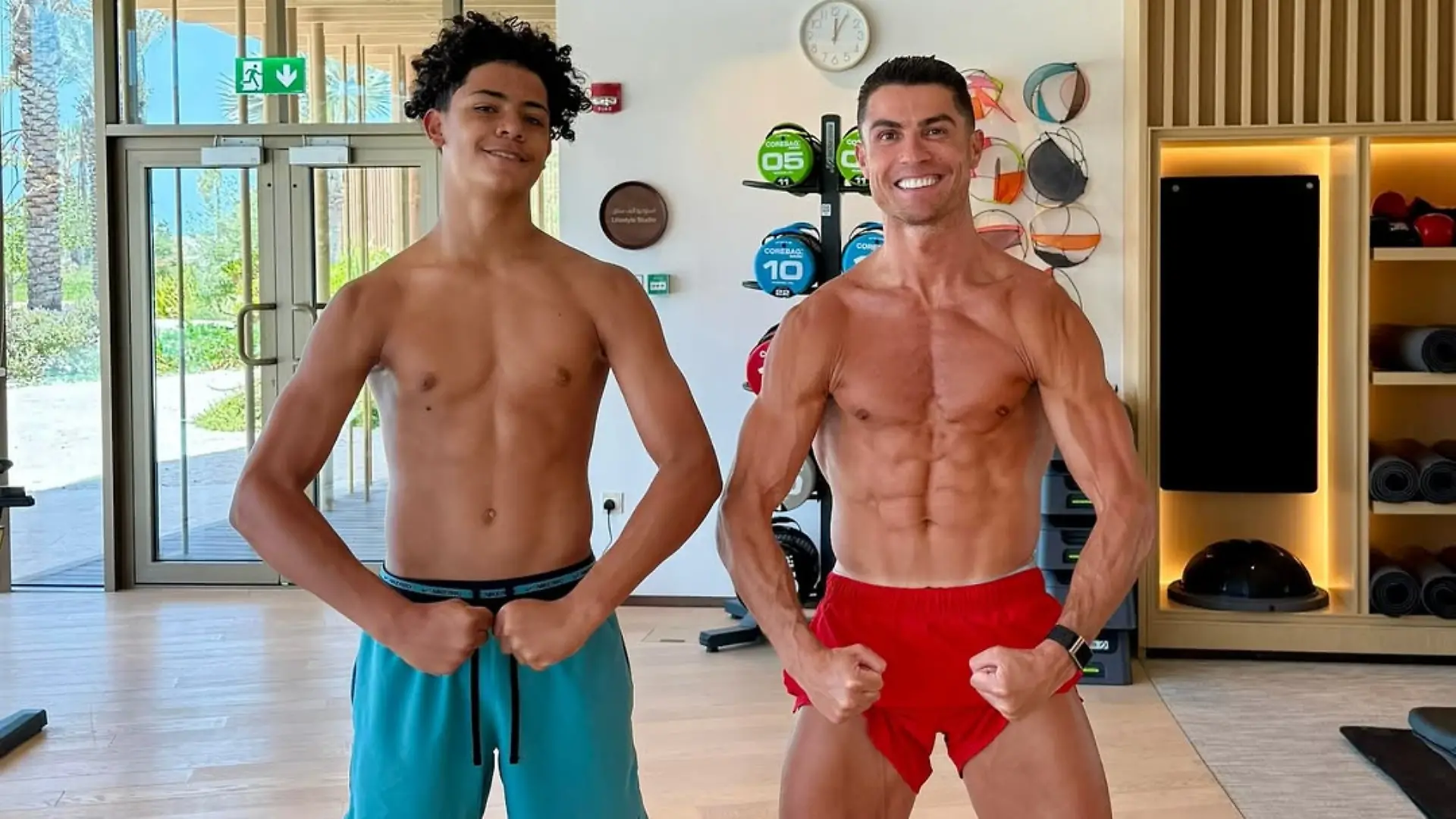 Cristiano Ronaldo und Cristiano Ronaldo jr. stehen nur mir Sportshorts bekleidet in einem Fitnessstudio und zeigen ihre Muskeln