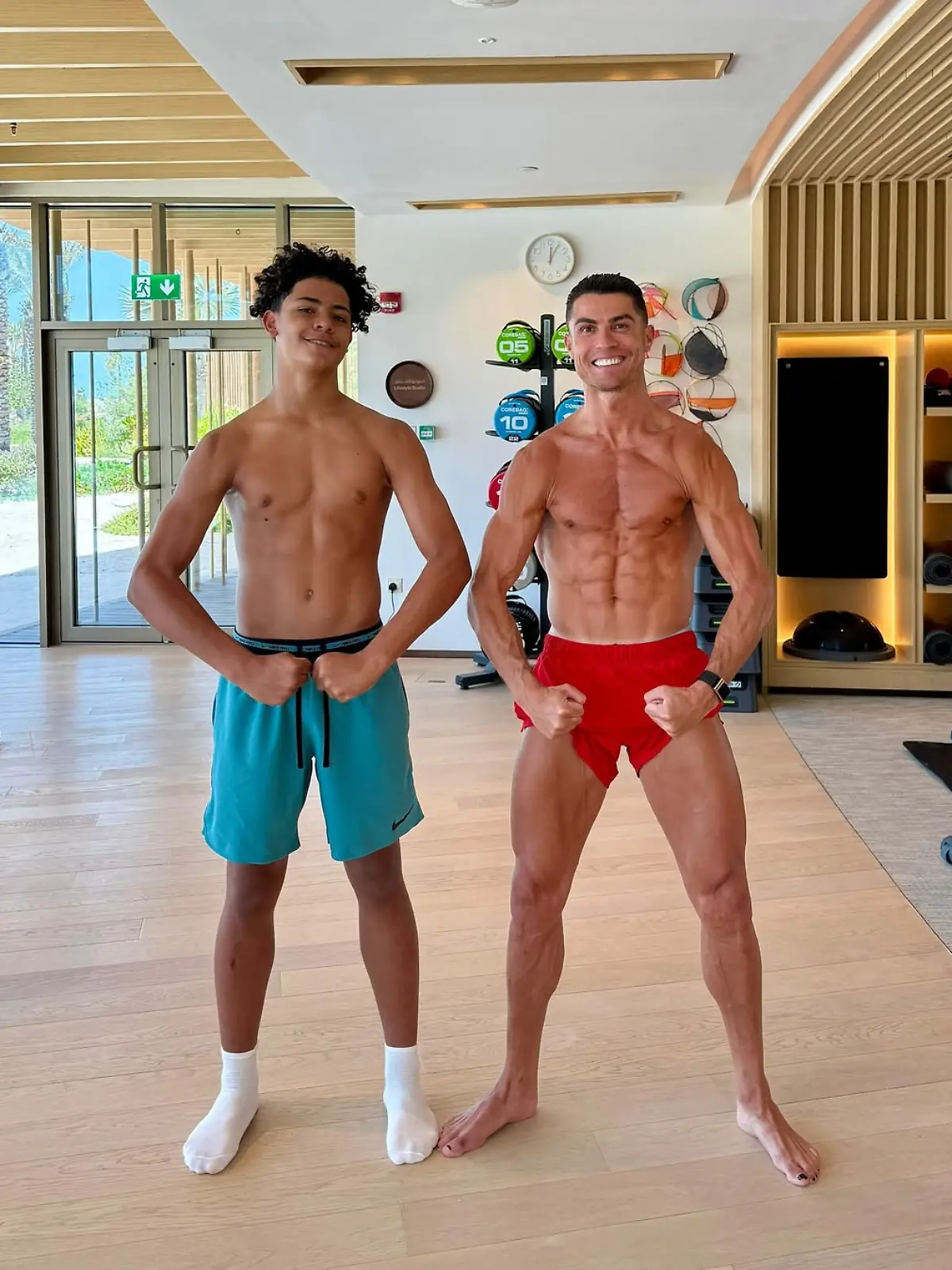 Cristiano Ronaldo und Cristiano Ronaldo jr. stehen nur mir Sportshorts bekleidet in einem Fitnessstudio und zeigen ihre Muskeln