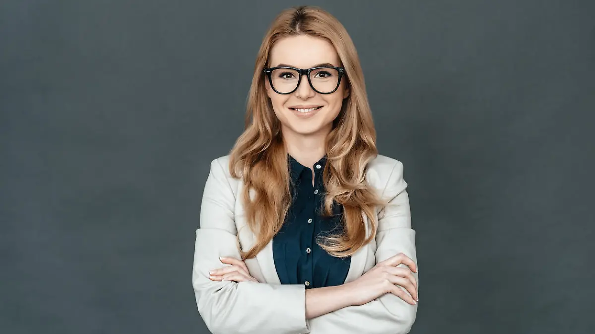 Gorgeous woman in smart casual wear keeping arms crossed and looking at camera while standing against grey background