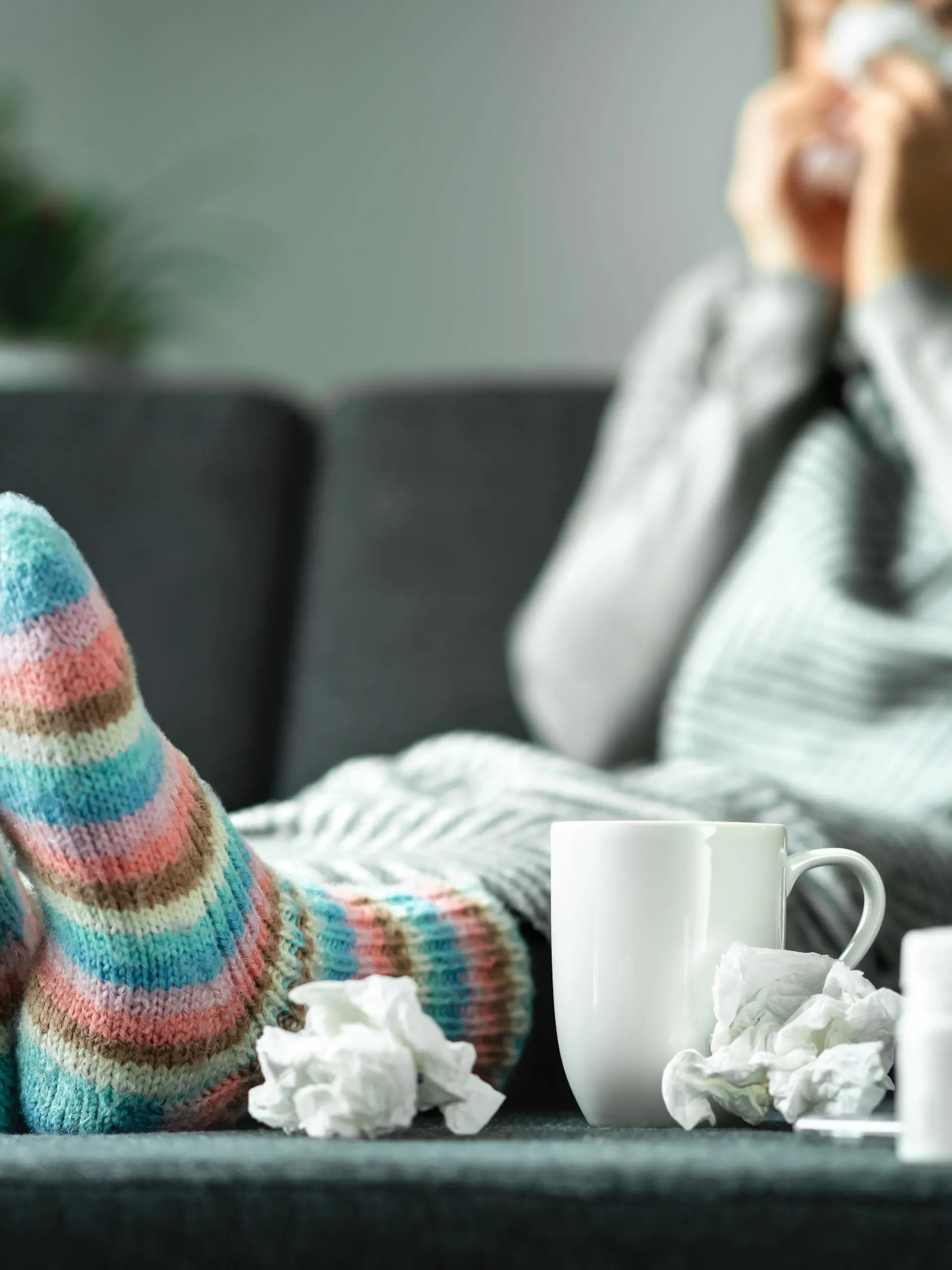 Sick woman with flu, cold, fever and cough sitting on couch at home. Ill person blowing nose and sneezing with tissue and handkerchief. Woolen socks and medicine. Infection in winter. Resting on sofa.