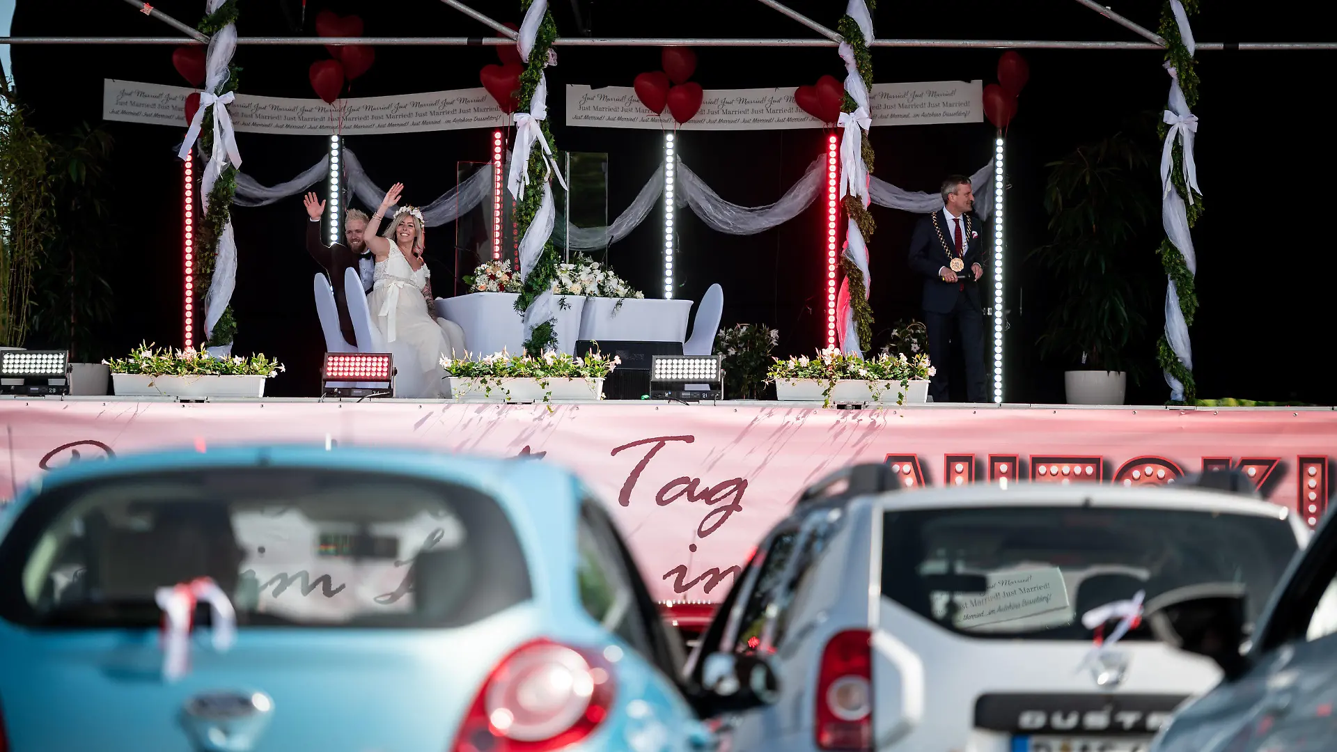 05.05.2020, Nordrhein-Westfalen, Düsseldorf: Das Brautpaar Janine und Philip Scholz (l) sitzt mit Düsseldorfs Oberbürgermeister Thomas Geisel (SPD) bei ihrer Trauung im Autokino auf der Bühne. Drei Paare geben sich im Autokino das Ja-Wort. Ihre Gäste können die Trauung über das Autoradio mithören. Das Paar heiratete ganz in rot-weiß · zu Ehren des 125. Jubiläums des Fußballclubs Fortuna Düsseldorf. Foto: Fabian Strauch/dpa +++ dpa-Bildfunk +++