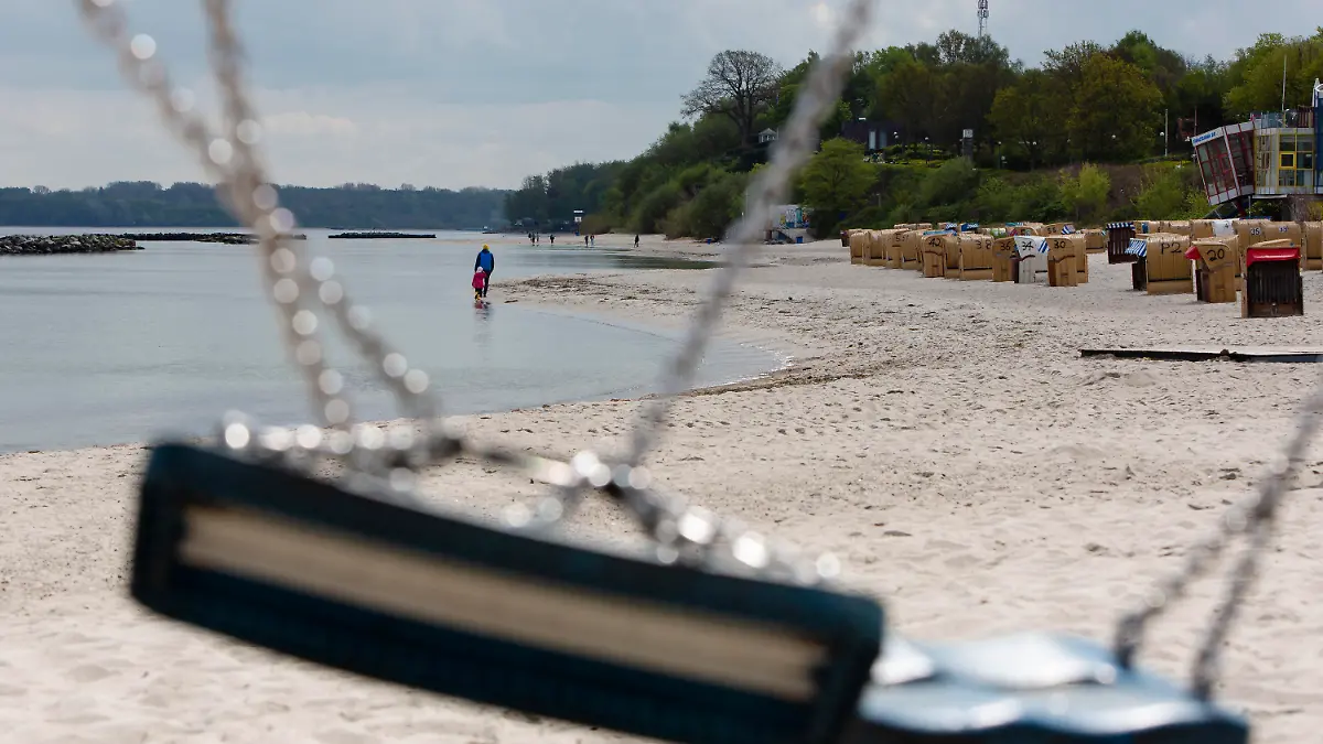 01.05.2020, Schleswig-Holstein, Kiel: Zwei Personen sind bei Sonnenschein durch eine zusammengeknotete Schaukel während ihres Spaziergangs am Strand von Kiel-Schilksee zu sehen. Sonne und Regen wechselten sich am 1. Mai 2020 in Schleswig-Holstein ab. Foto: Frank Molter/dpa +++ dpa-Bildfunk +++