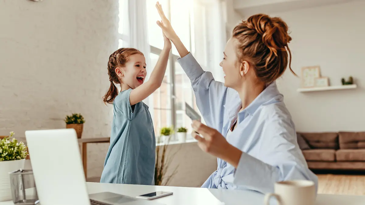 Low angle of excited small daughter giving high five to mother and screaming while celebrating successful online shopping using laptop at table in light living room