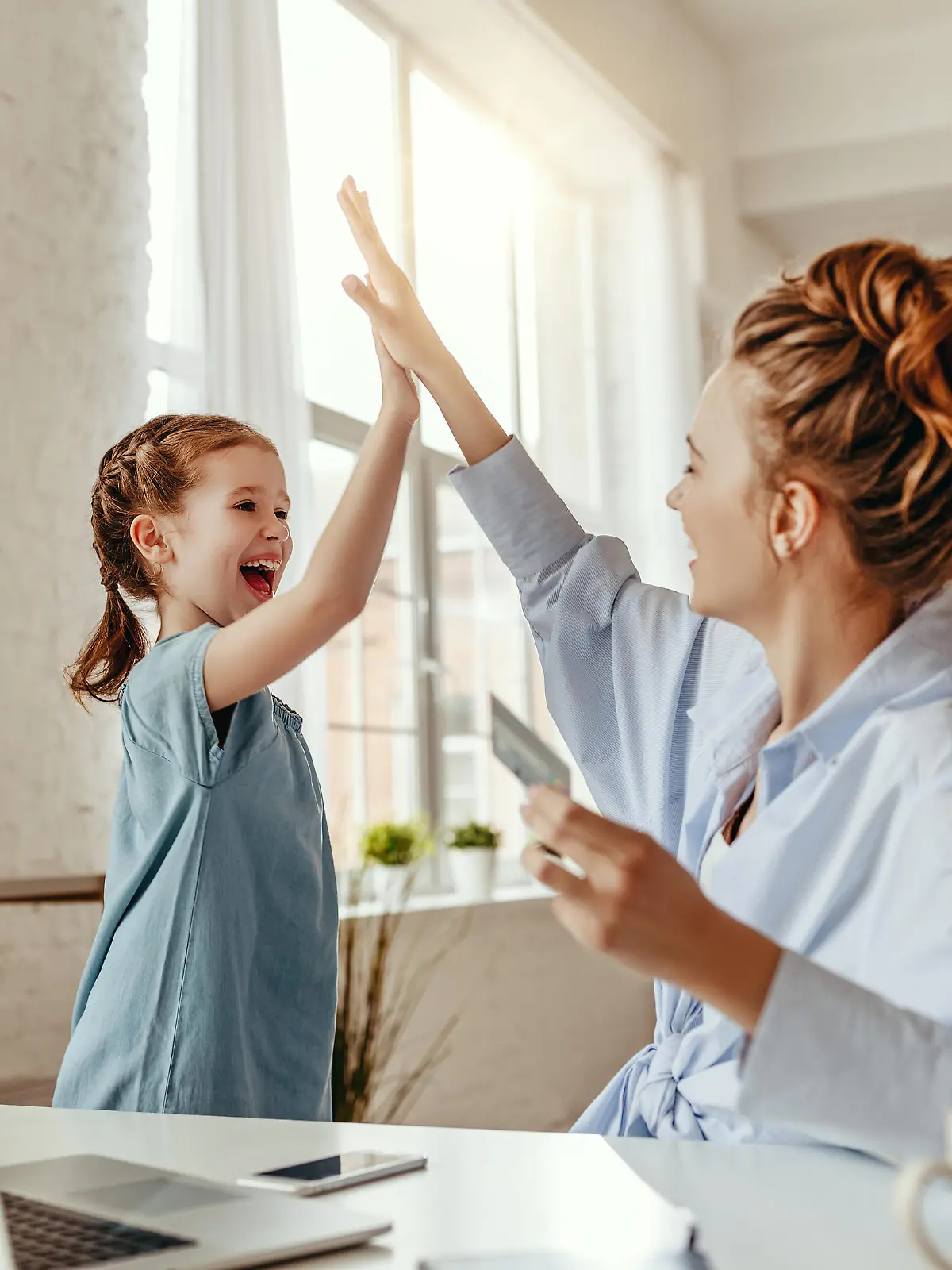 Low angle of excited small daughter giving high five to mother and screaming while celebrating successful online shopping using laptop at table in light living room