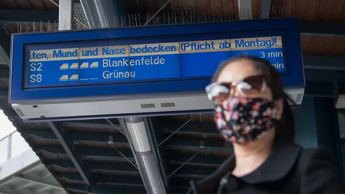 25.04.2020, Berlin: "Mund und Nase bedecken (Pflicht ab Montag!)" ist auf einer elektronischen Anzeigetafel an der S-Bahn-Station Pankow-Heinersdorf zu lesen, während eine Frau, die einen Mundschutz trägt, auf die S-Bahn wartet. Ab dem 27. April 2020 ist ein Mundschutz bei der Nutzung des öffentlichen Nahverkehrs in Berlin verpflichtend. Foto: Jörg Carstensen/dpa +++ dpa-Bildfunk +++