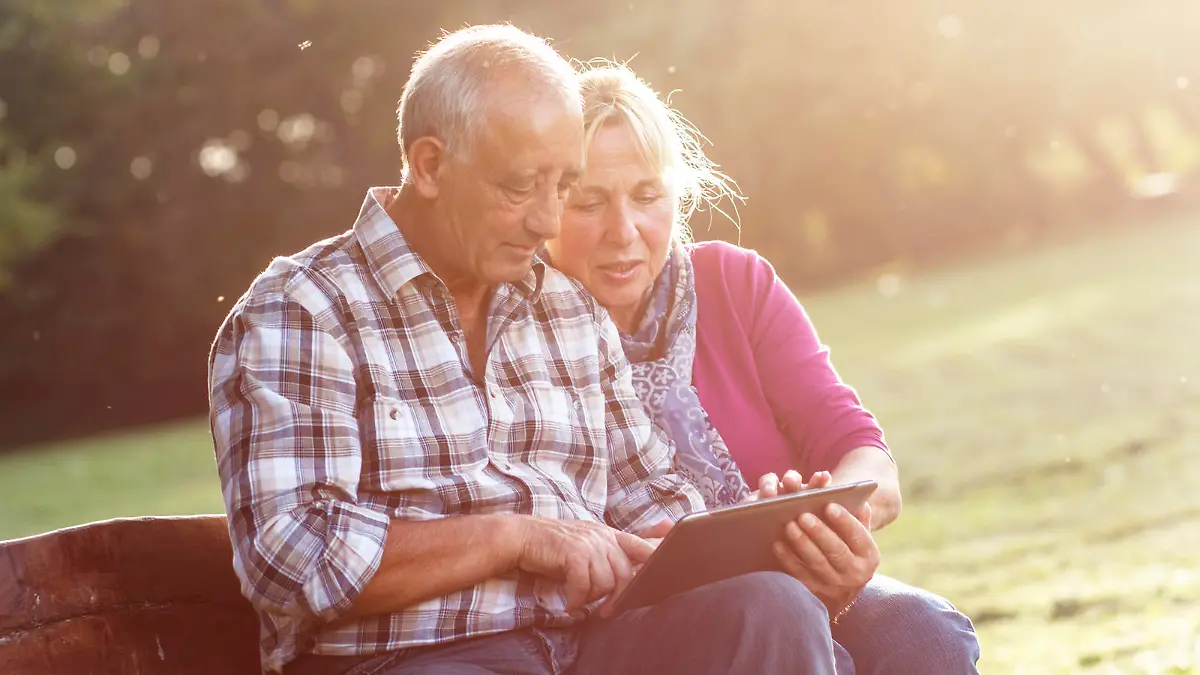 Senior couple sitting on a park bench on sunny autumn day with tablet and relaxing.