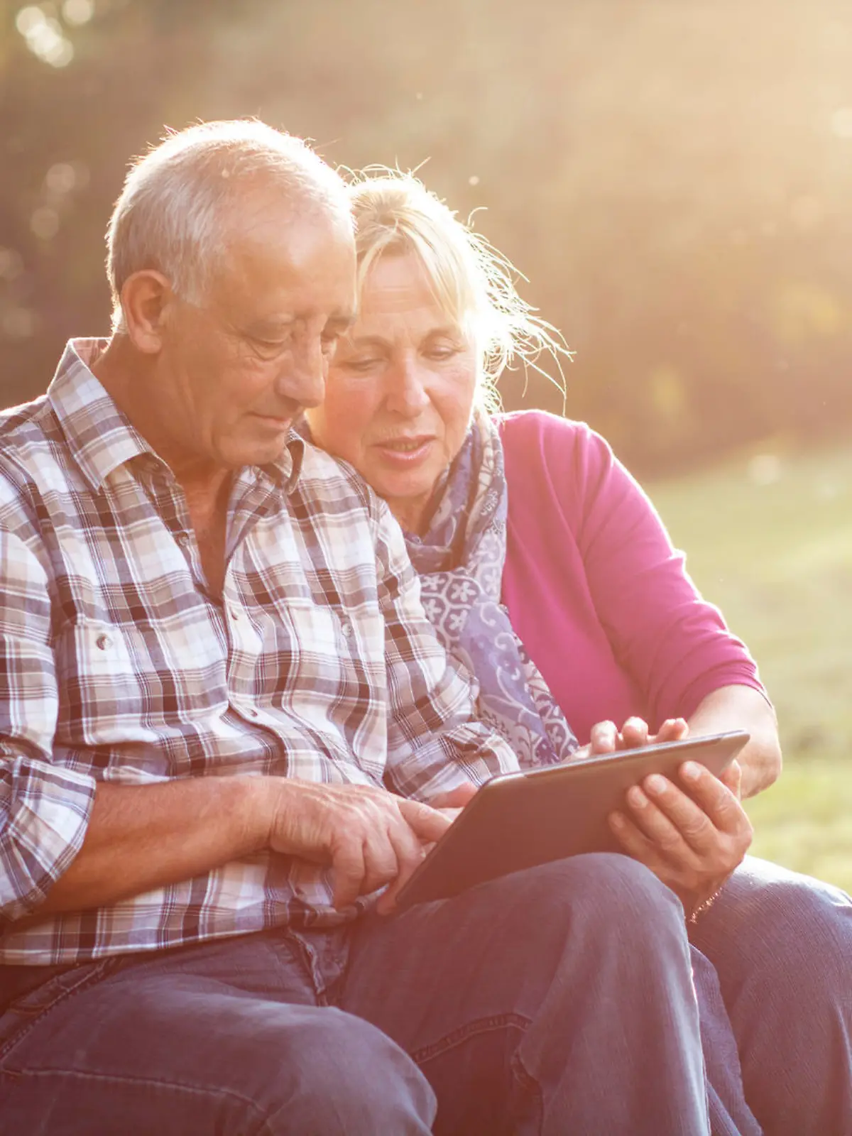 Senior couple sitting on a park bench on sunny autumn day with tablet and relaxing.