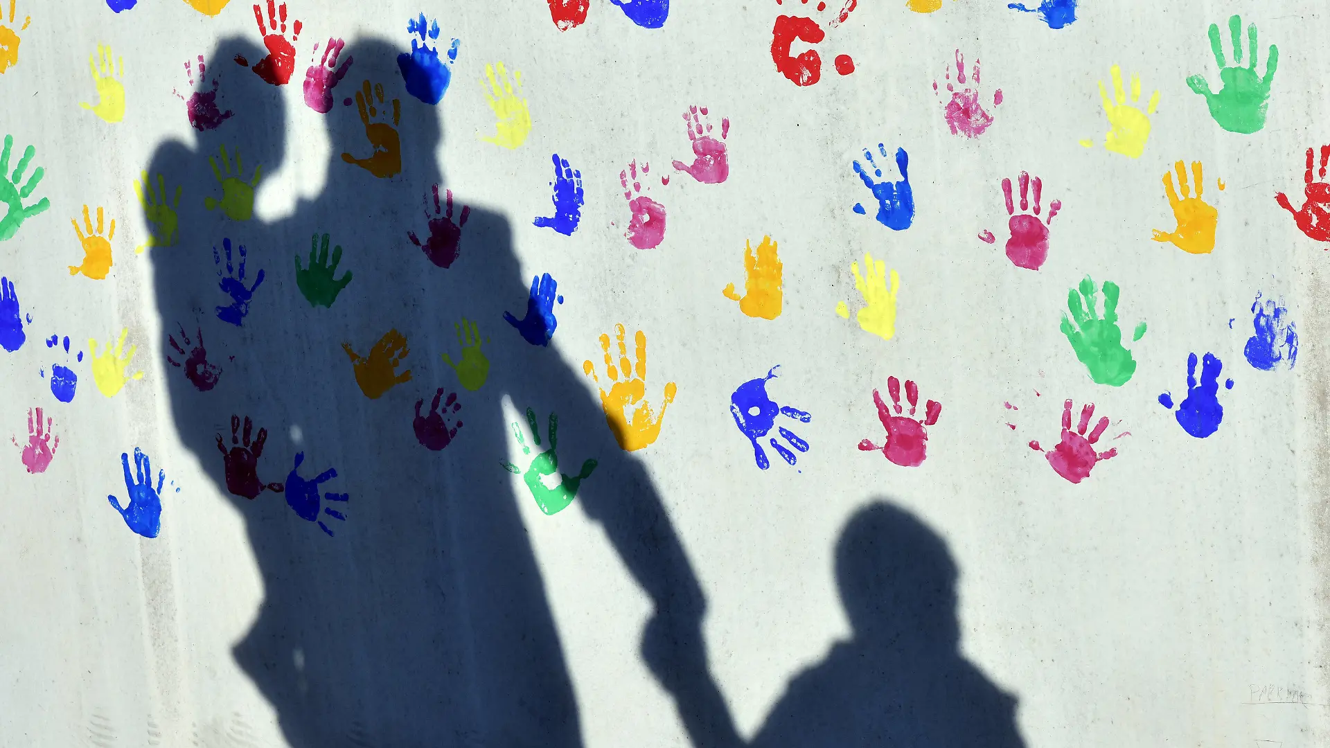 ILLUSTRATION - 08.04.2015, Bayern, München: Ein Mann mit einem Kind auf dem Arm und einem an der Hand wirft einen Schatten auf eine mit bunten Handabdrücken bemalte Wand einer Kindertagesstätte. (zu "Zahl der Pflegeeltern in Brandenburg") Foto: Peter Kneffel/dpa +++ dpa-Bildfunk +++