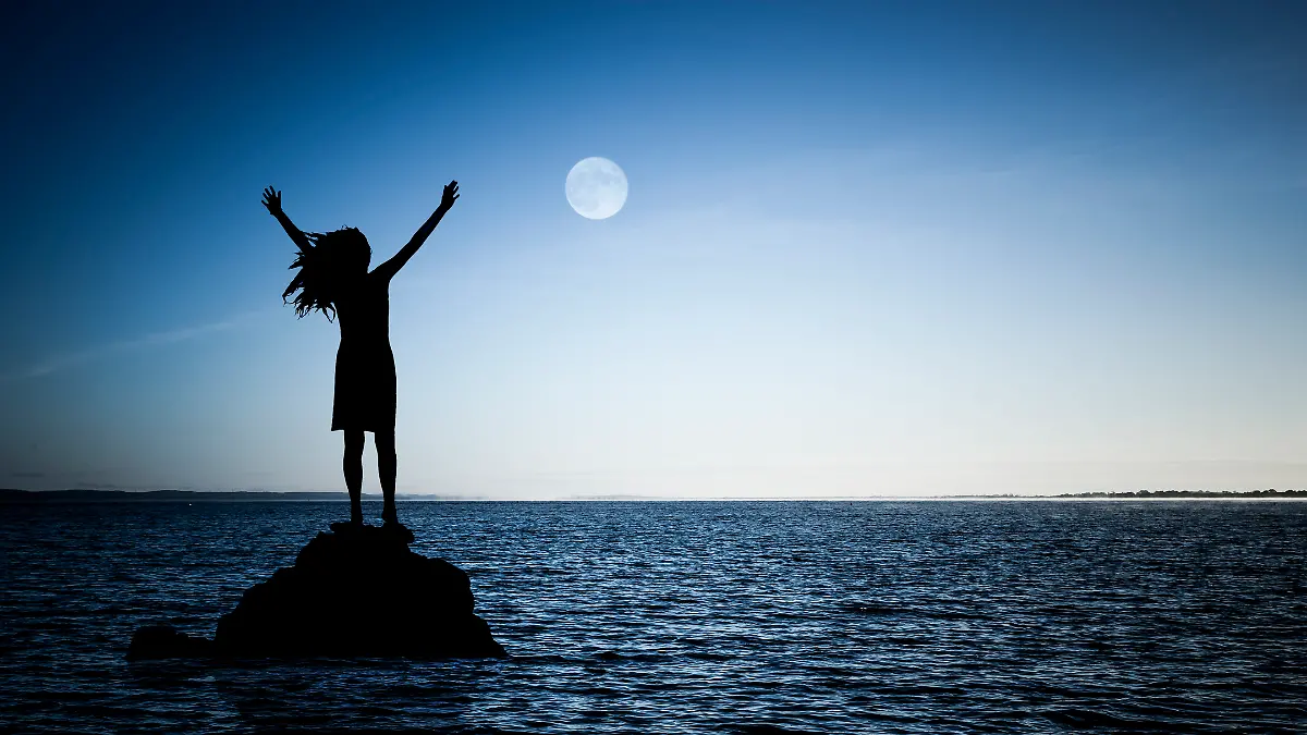 Eine Frau steht auf einem Felsen im Wasser und betet den Vollmond an.