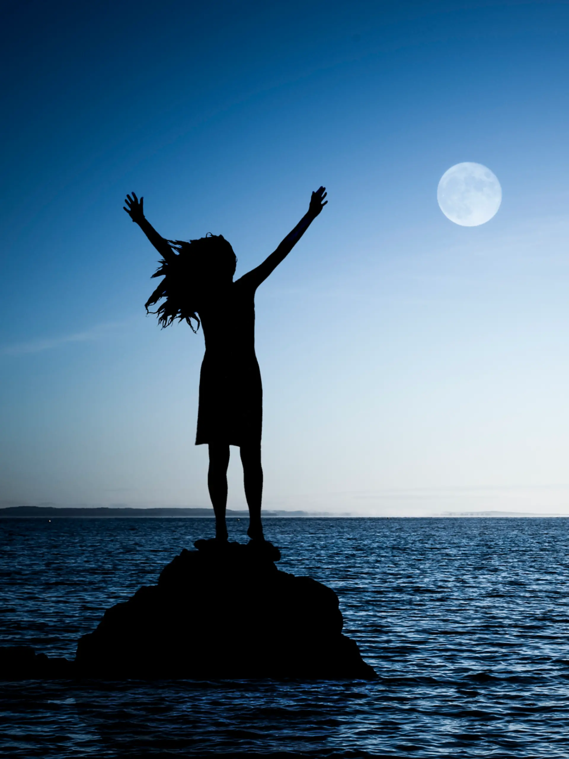 Eine Frau steht auf einem Felsen im Wasser und betet den Vollmond an. 