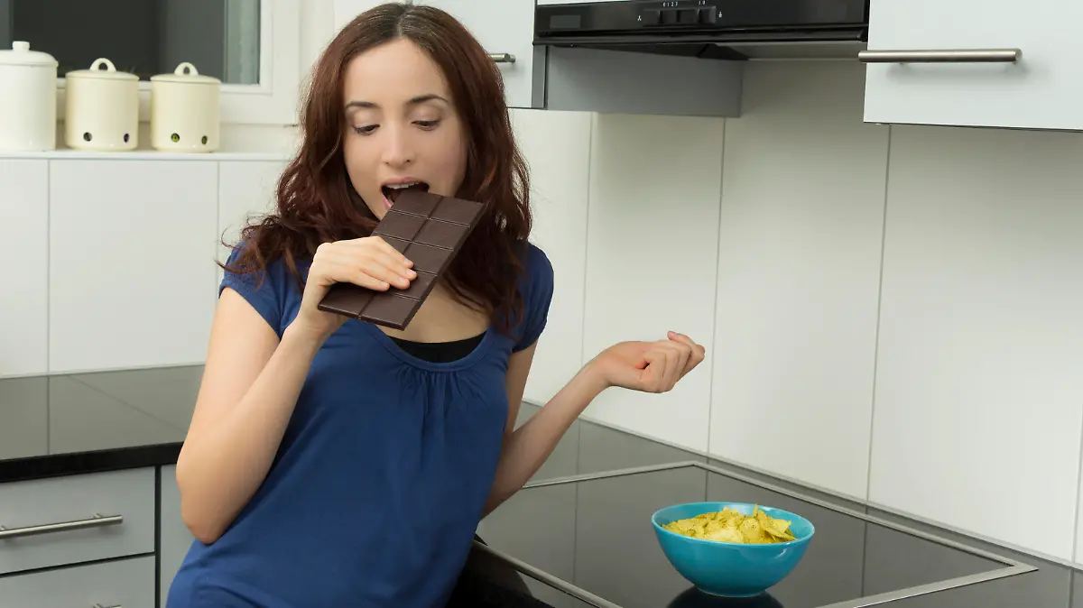 Junge Frau steht in der Küche und beißt in eine Tafel Bitterschokolade. Auf der Küchenzeile steht eine Schüssel mit Kartoffelchips.