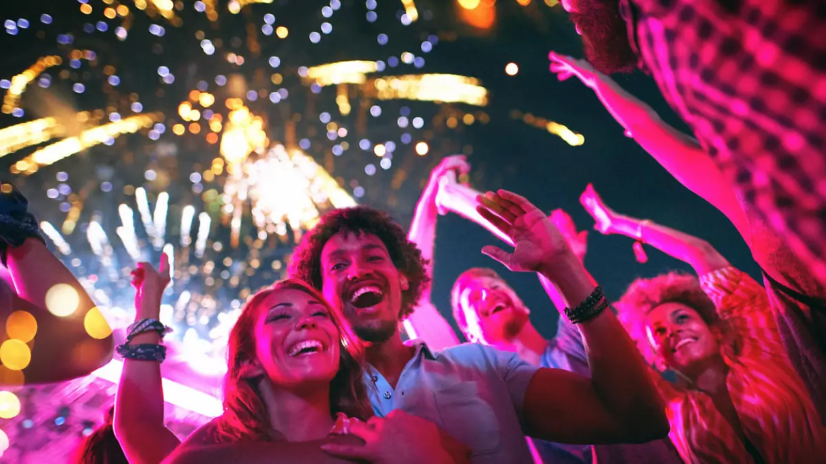Closeup low angle view of group of multi ethnic young adults dancing and enjoying an open air concert on a summer afternoon. They are standing in a circle while fireworks popping in the background.