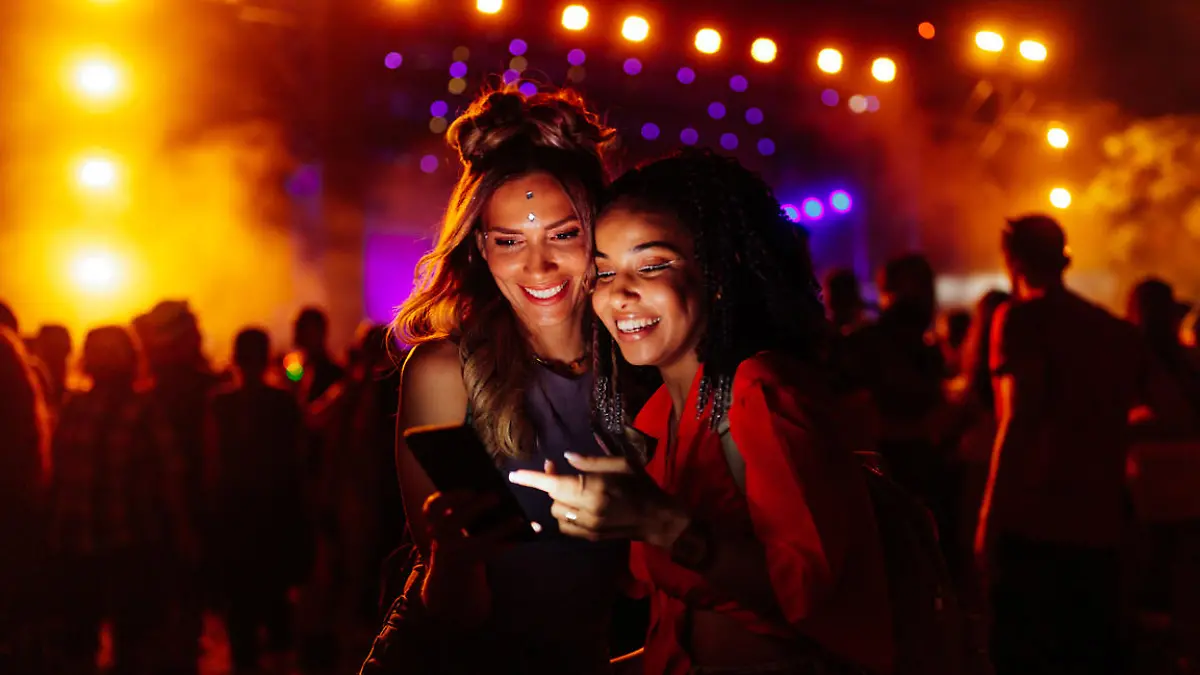 Two biracial women in a crowd at a concert using cellphone together