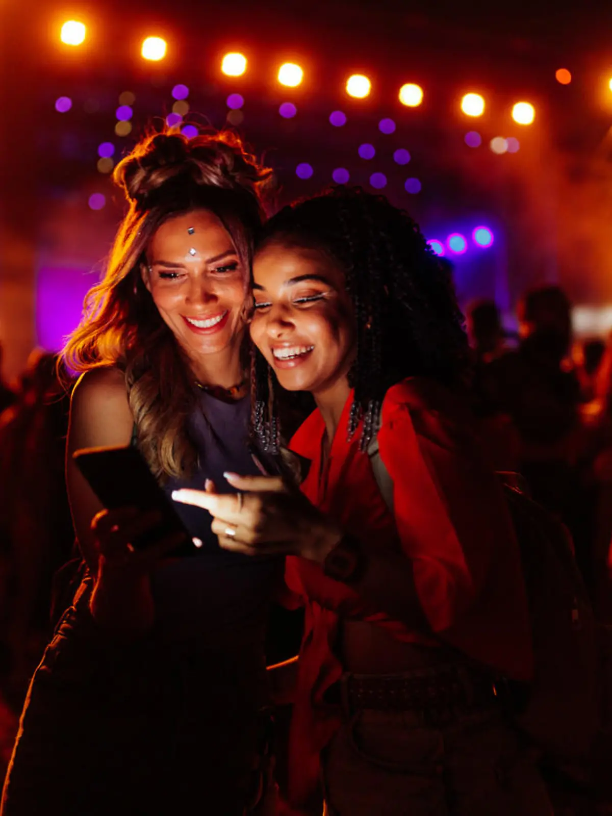 Two biracial women in a crowd at a concert using cellphone together