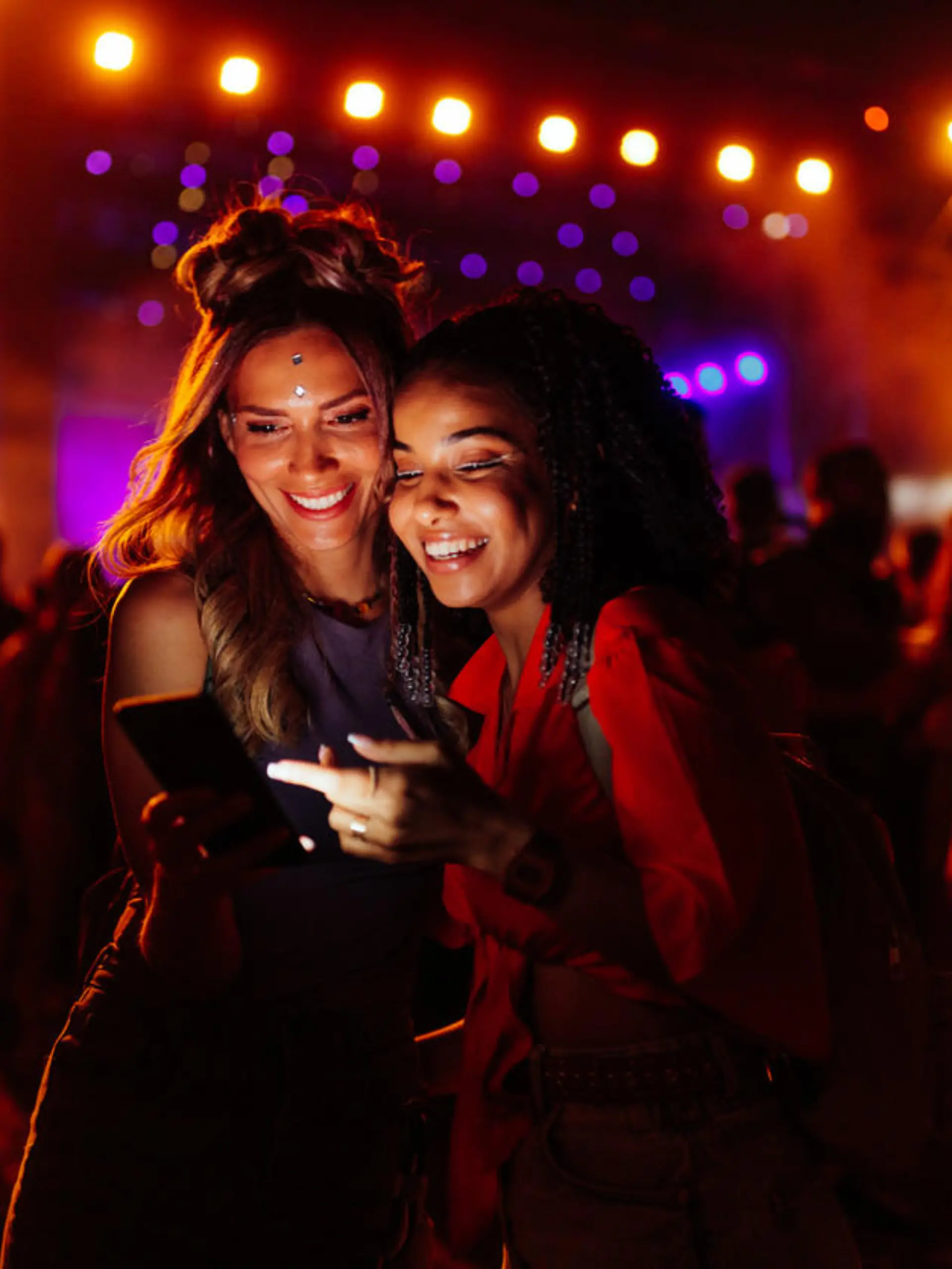 Two biracial women in a crowd at a concert using cellphone together