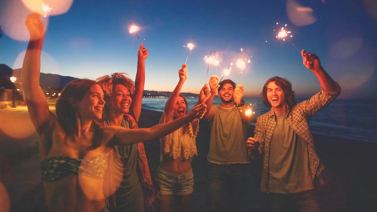 Group of friends playing with sparklers and fireworks on the beach at sunset. They are dancing and having fun . All are happy and smiling and laughing.