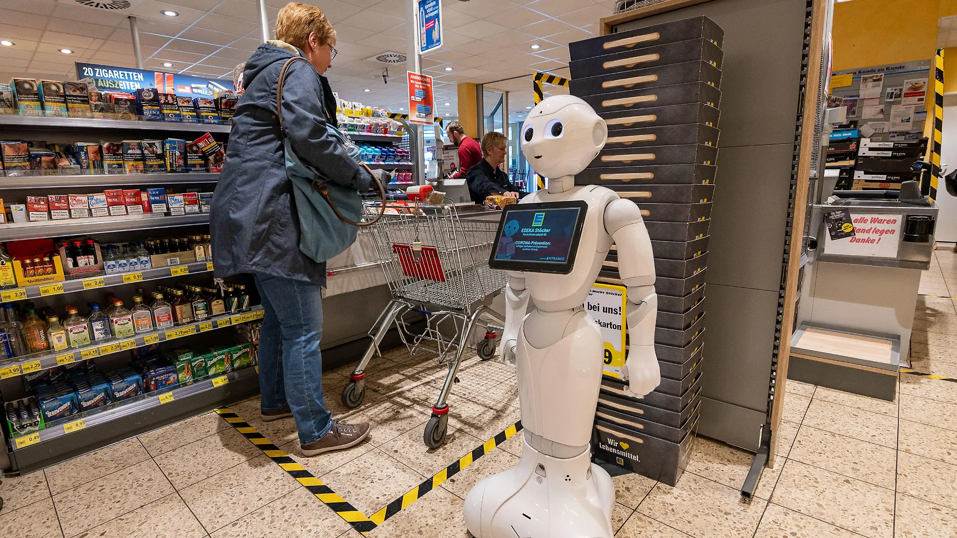 28.03.2020, Nordrhein-Westfalen, Lindlar: Pflegeroboter «Pepper» steht im Kassenbereich des Edeka-Marktes und weist Kunden darauf hin, Abstand zu halten und sich vor einer Ansteckung mit dem Coronavirus zu schützen. Außerdem wirbt er für Rücksichtnahme und Solidarität untereinander und rät mit kindlich-freundlichem «Gesichtsausdruck» von Hamsterkäufen ab. (zu dpa "Abstand wahren: Roboter spricht Kunden in Kassenzone an") Foto: Markus Klümper/dpa +++ dpa-Bildfunk +++