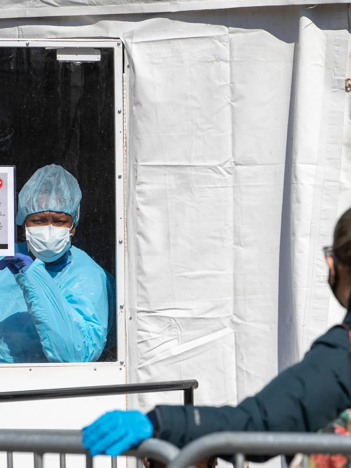 26.03.2020, USA, New York: Eine medizinische Mitarbeiterin des Krankenhauses «The Brooklyn Hospital Center» steht auf einem Covid-19-Testgelände hinter einer Scheibe eines Zelts, und ruft den nächsten Patienten der Warteschlange auf. In den USA gibt es inzwischen laut den offiziellen Angaben weltweit die meisten Corona-Infizierten. Am schlimmsten betroffen ist New York. Foto: Mary Altaffer/AP/dpa +++ dpa-Bildfunk +++