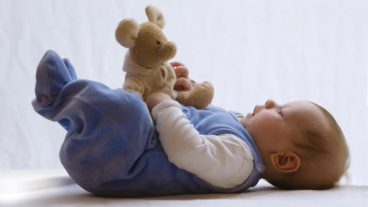 Baby liegt im Strampler auf dem Bett und spielt mit Stofftier
