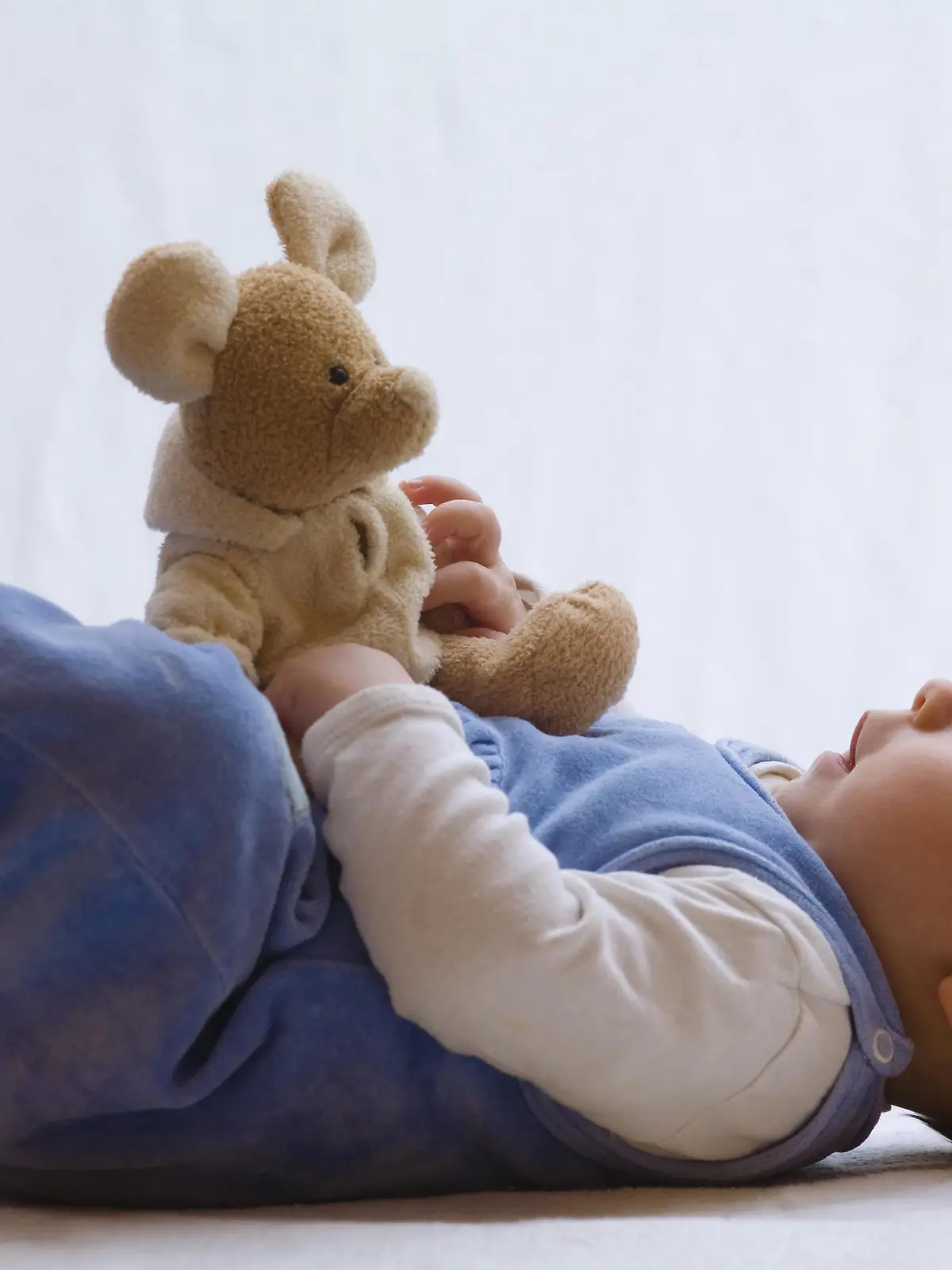 Baby liegt im Strampler auf dem Bett und spielt mit Stofftier