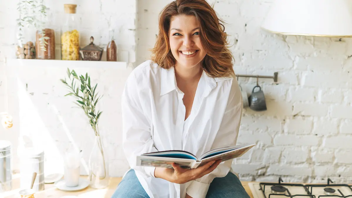 Gorgeous happy young woman plus size body positive in blue jeans and white shirt reading cooking book in home kitchen