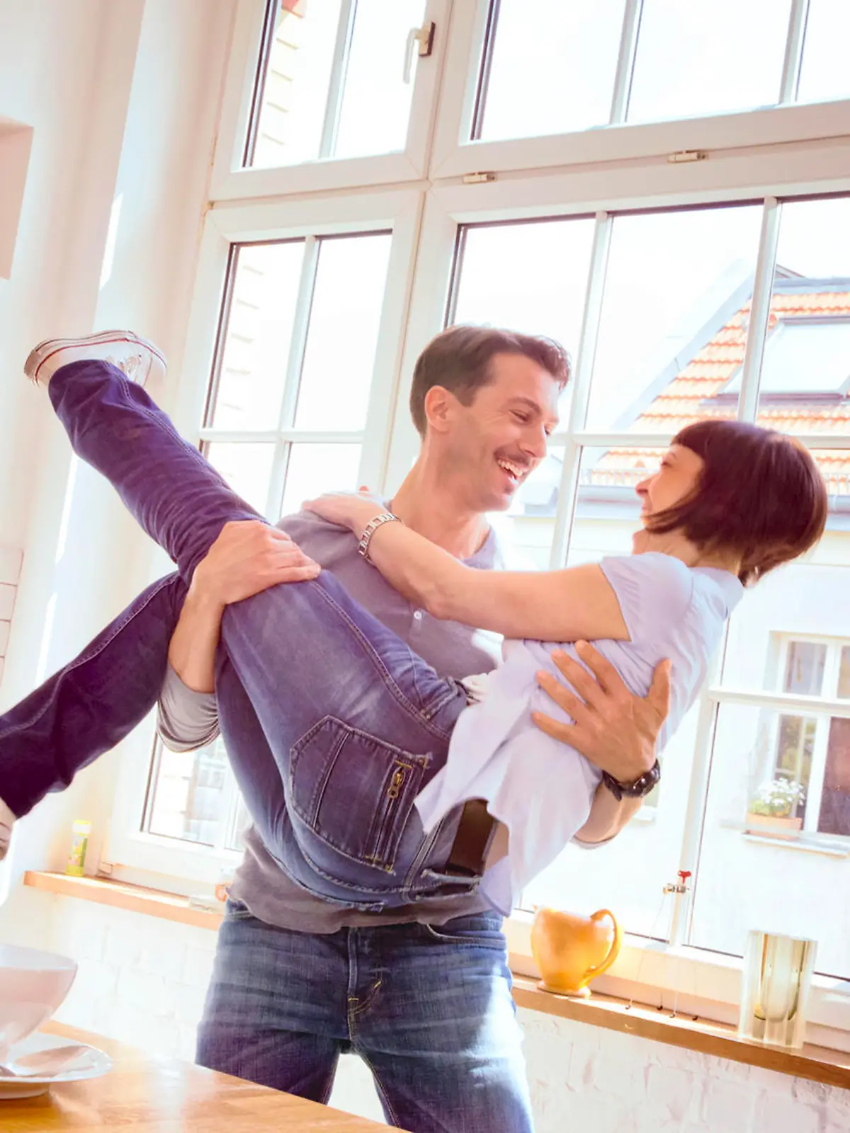 Young couple enjoying their love in the kitchen one early morning. The man has lifted his lover in his arm and they're both laughing.