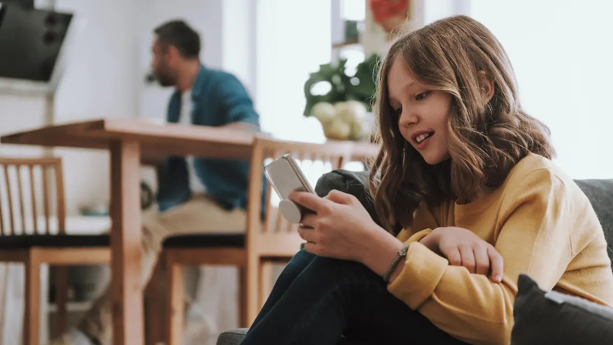 Portrait of adorable child sitting on couch and surfing the internet on smartphone. Her father on blurred background