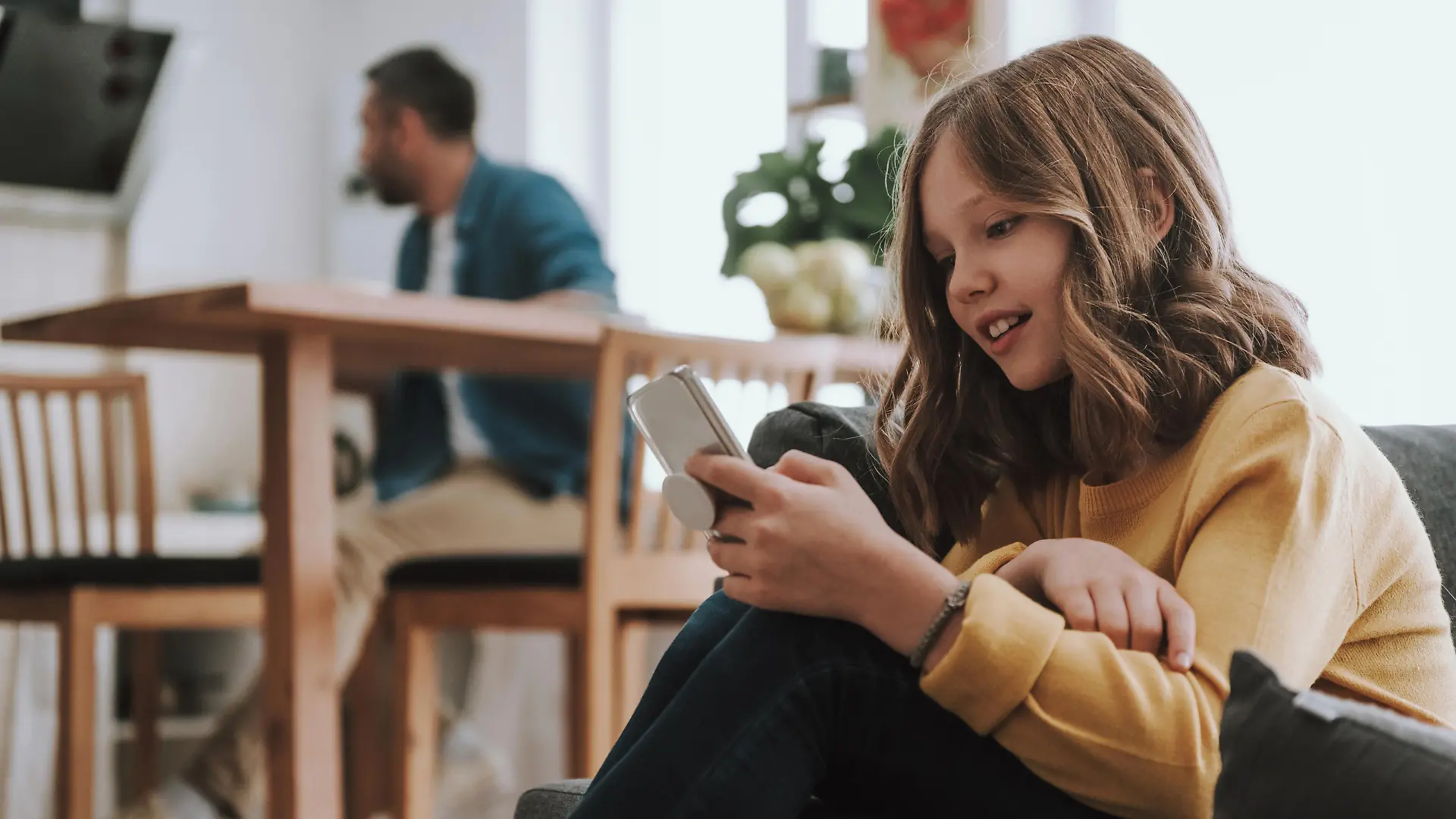 Portrait of adorable child sitting on couch and surfing the internet on smartphone. Her father on blurred background