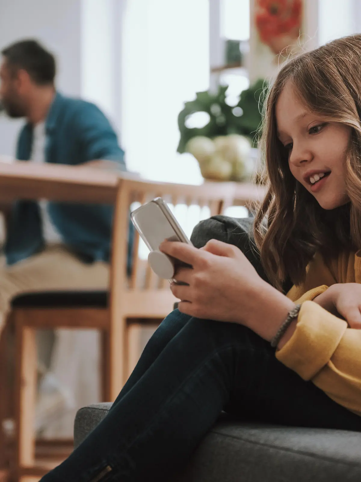 Portrait of adorable child sitting on couch and surfing the internet on smartphone. Her father on blurred background