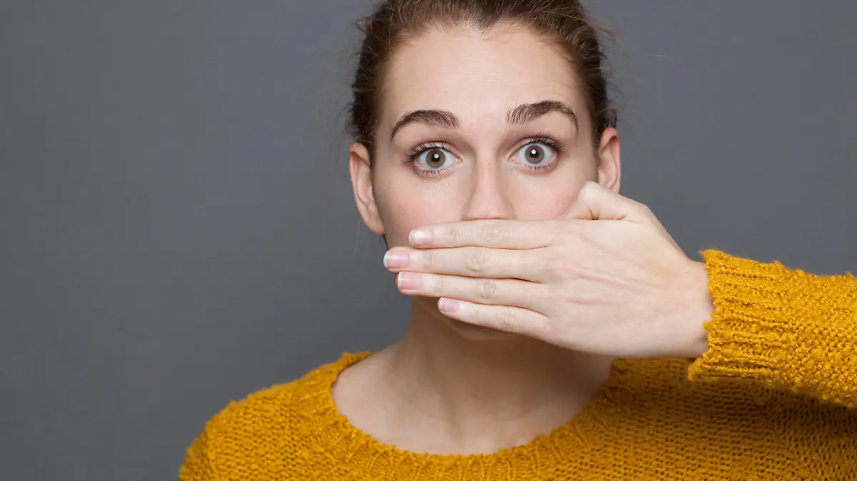 negative feelings concept - portrait of amazed beautiful 20s girl covering her mouth for gag metaphor,studio shot on gray background