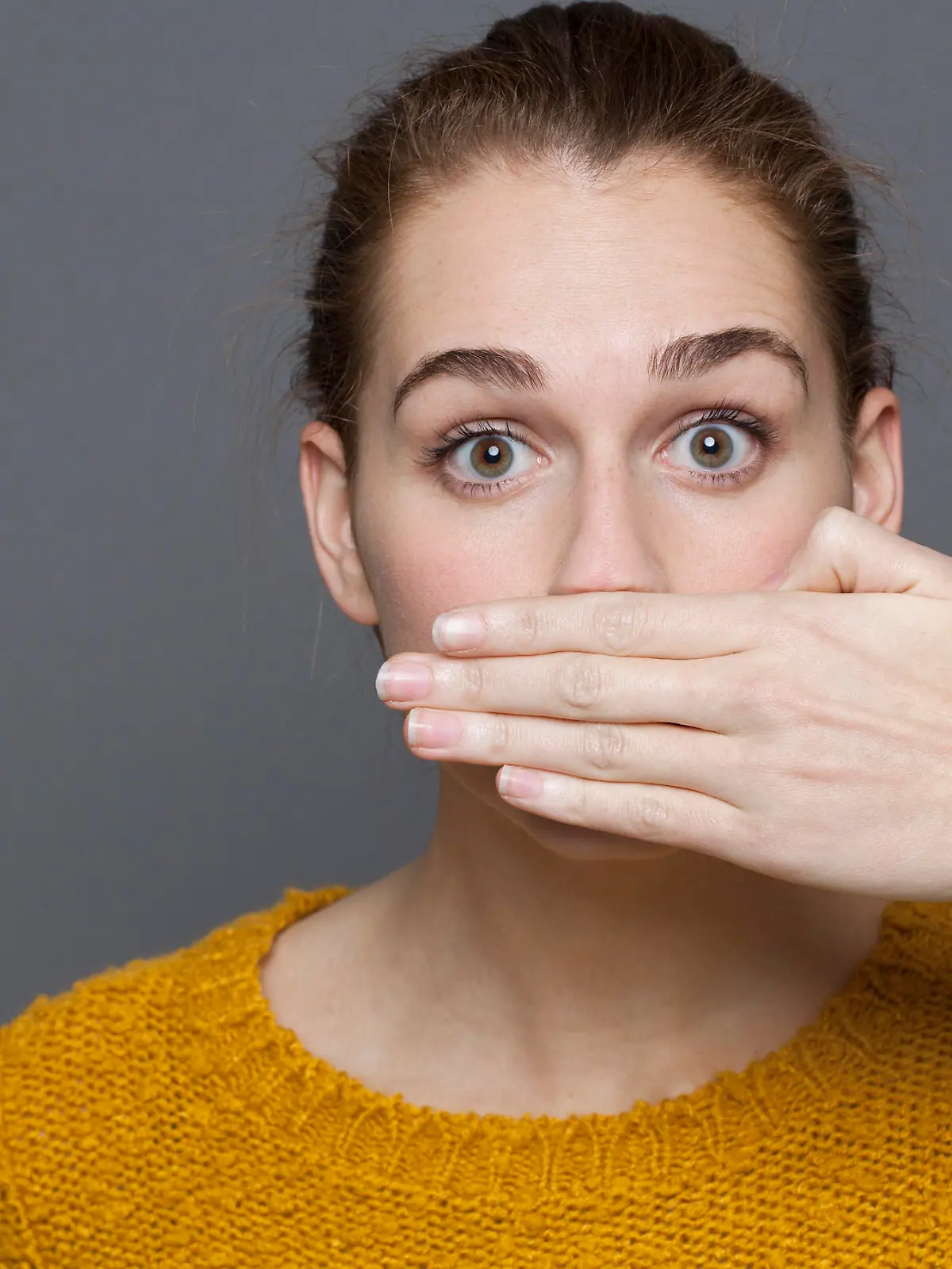 negative feelings concept - portrait of amazed beautiful 20s girl covering her mouth for gag metaphor,studio shot on gray background