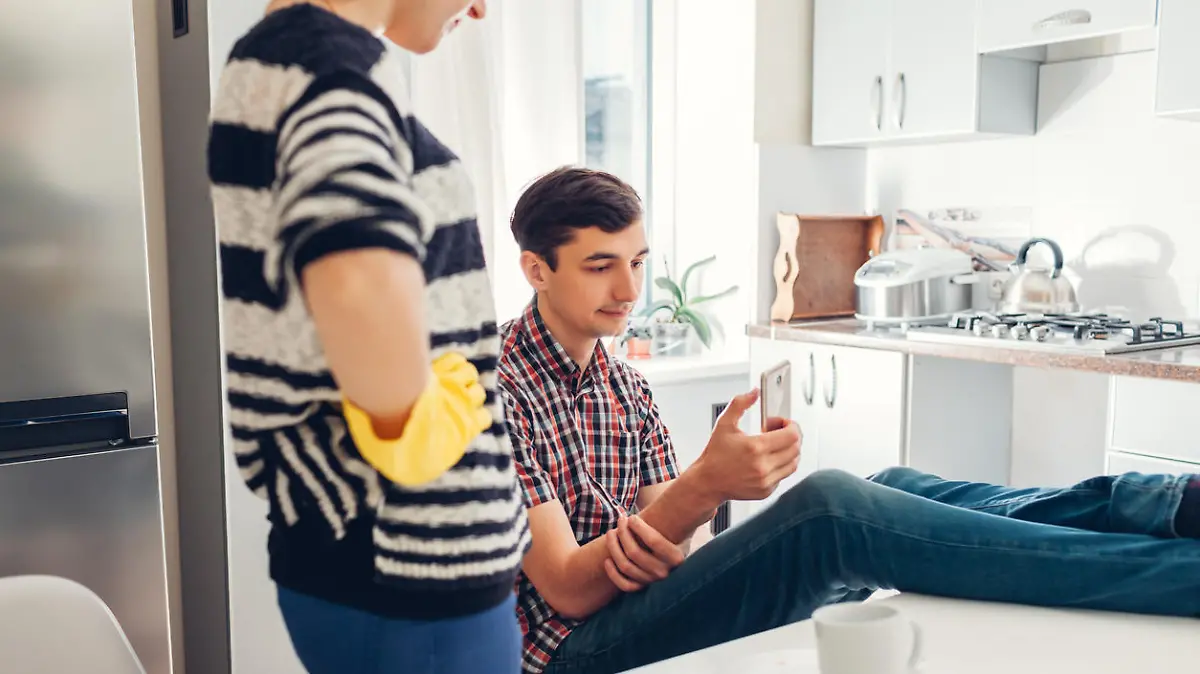 Lazy boyfriend hanging in phone while his girlfriend cleaning his dishes after dinner in kitchen. Inequality of men's and women's rights. Family conflict