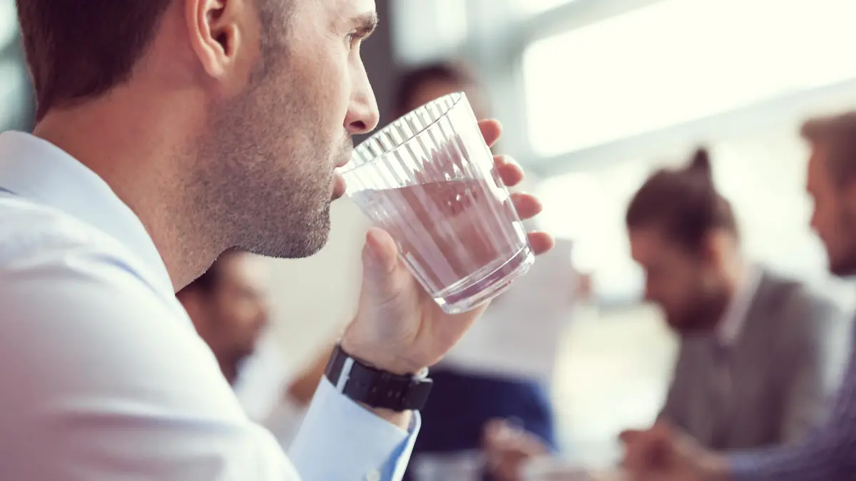 Close up of businessman drinking water at the meeting. In the background blured business team discussing in a board room in an office.
