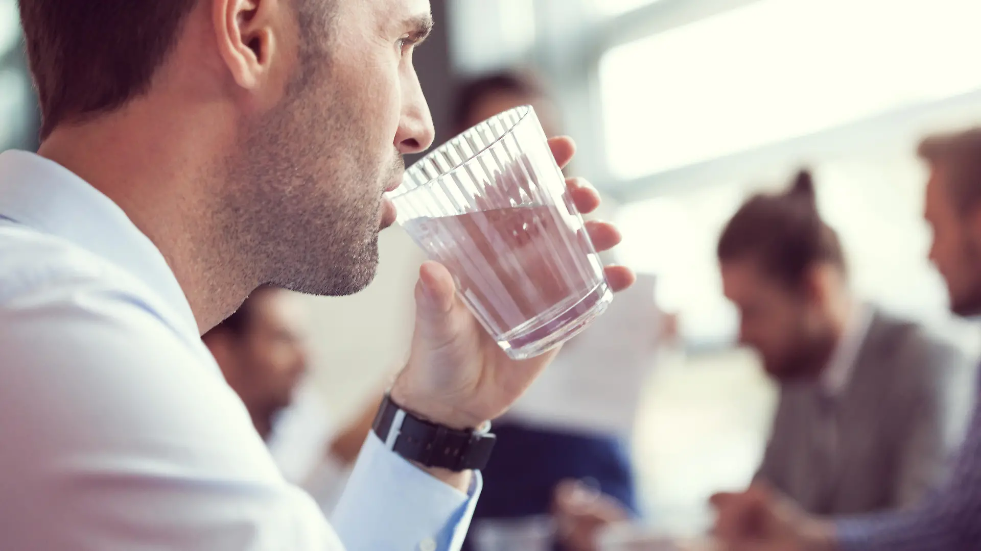 Close up of businessman drinking water at the meeting. In the background blured business team discussing in a board room in an office.