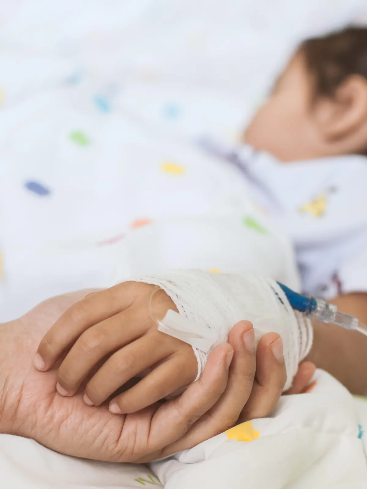 Mother hand holding sick daughter hand who have IV solution bandaged with love and care while she is sleeping on bed in the hospital