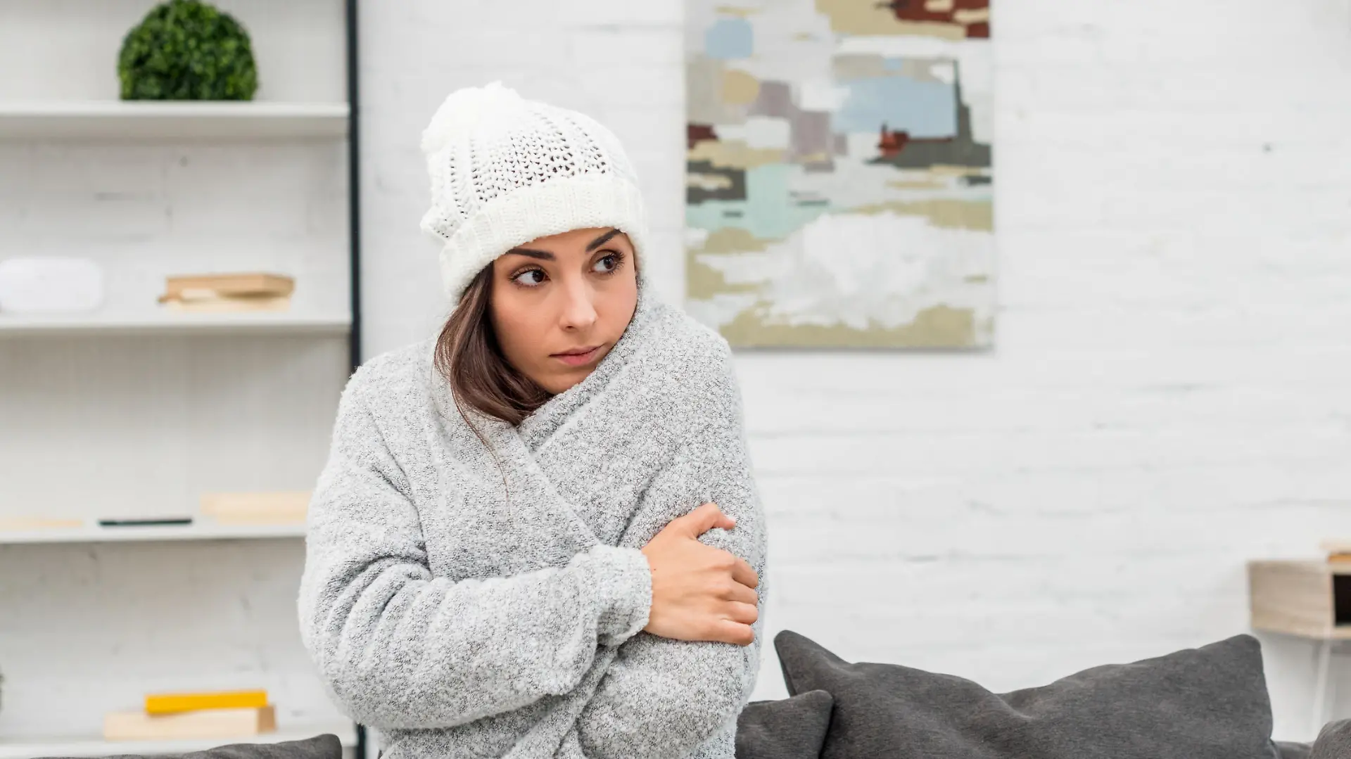 freezed young woman in warm clothes sitting on couch at home with air condition hanging on background